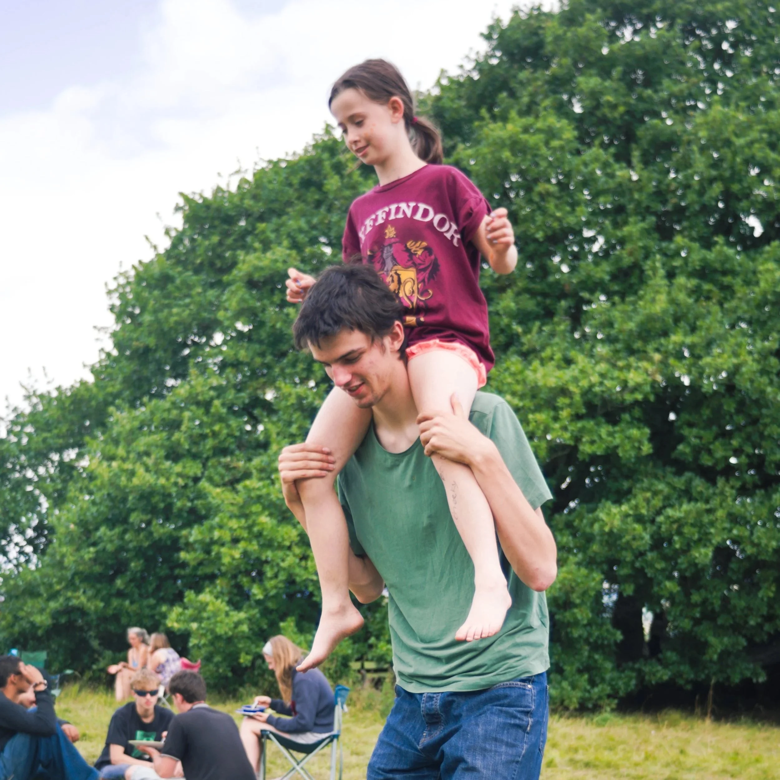 A young girl is sitting on the shoulders of a man at an outdoor gathering. The girl has brown hair tied back, is wearing a maroon shirt with the word 'Hufflepuff' and a crest on it, and is looking down and to her left. The man is wearing a green shirt, has short dark hair, and is smiling as he holds the girl steady. In the background, there are several people sitting on the grass, some with notebooks, and lush green trees under a partly cloudy sky.