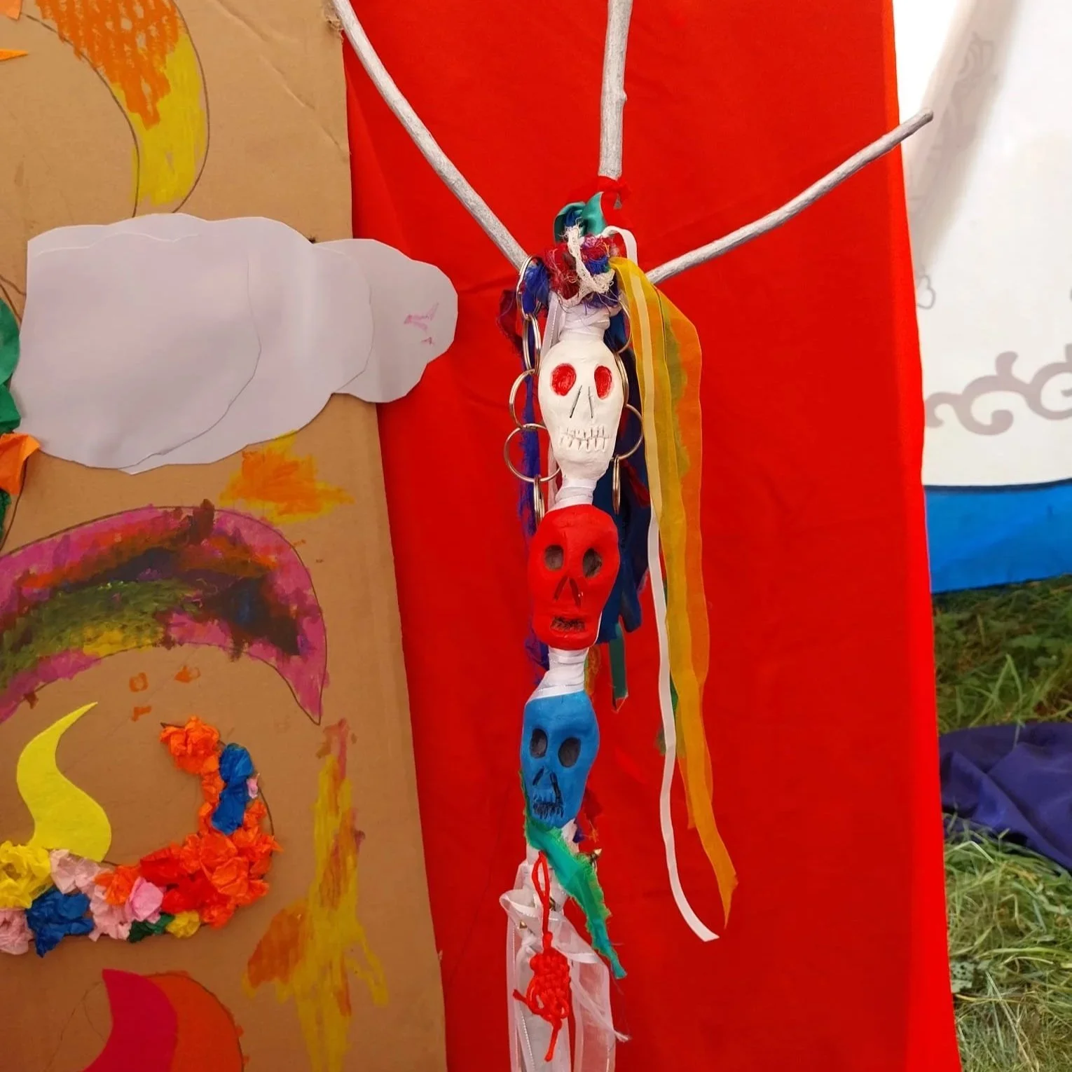 Decorative hanging craft with multiple colorful skulls made from paper or clay, strung vertically with ribbons and garlands, against a red fabric backdrop.