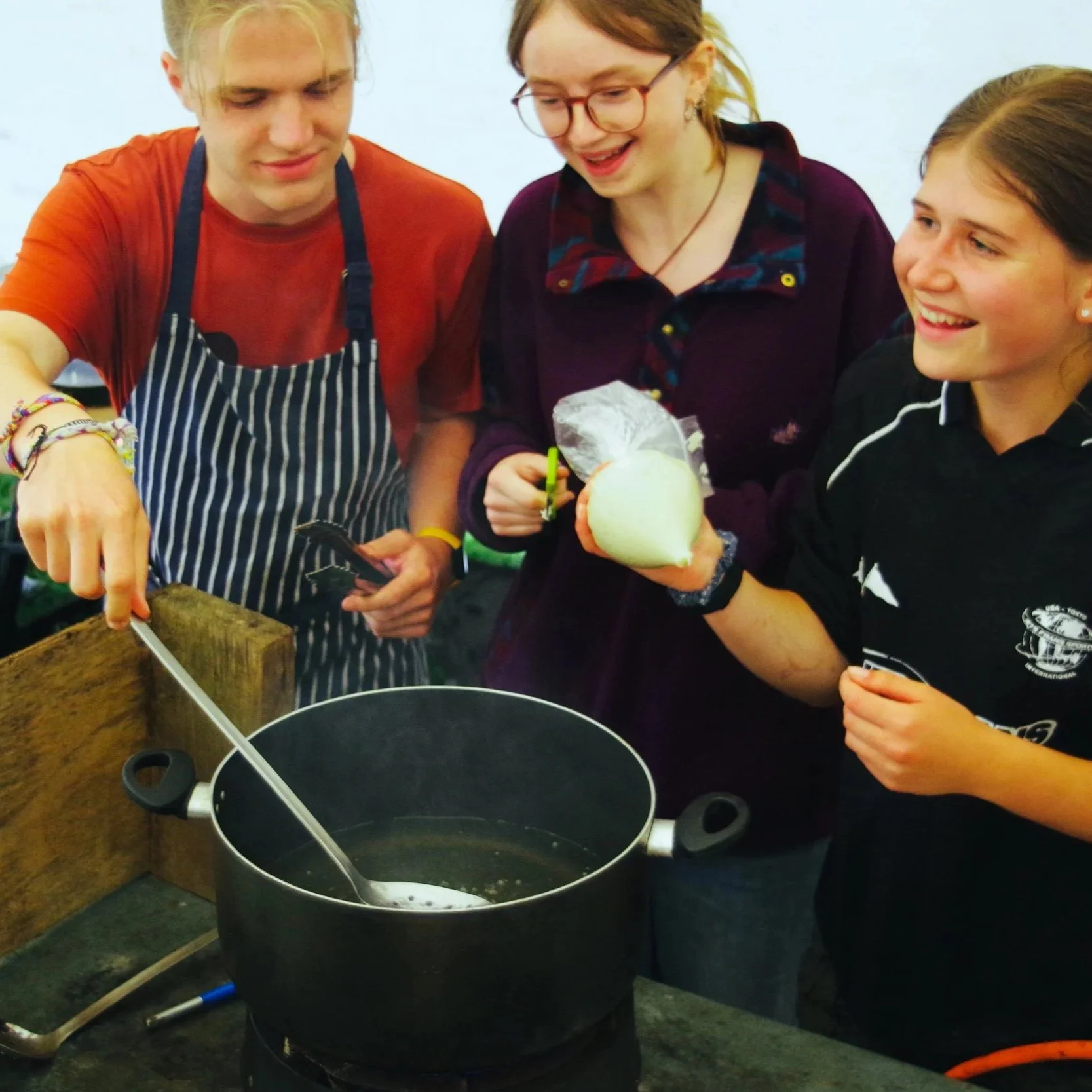 Three young people gather around a large black cooking pot, preparing food. One holds a peeled onion, while another is using a knife. They appear happy and engaged in cooking together.