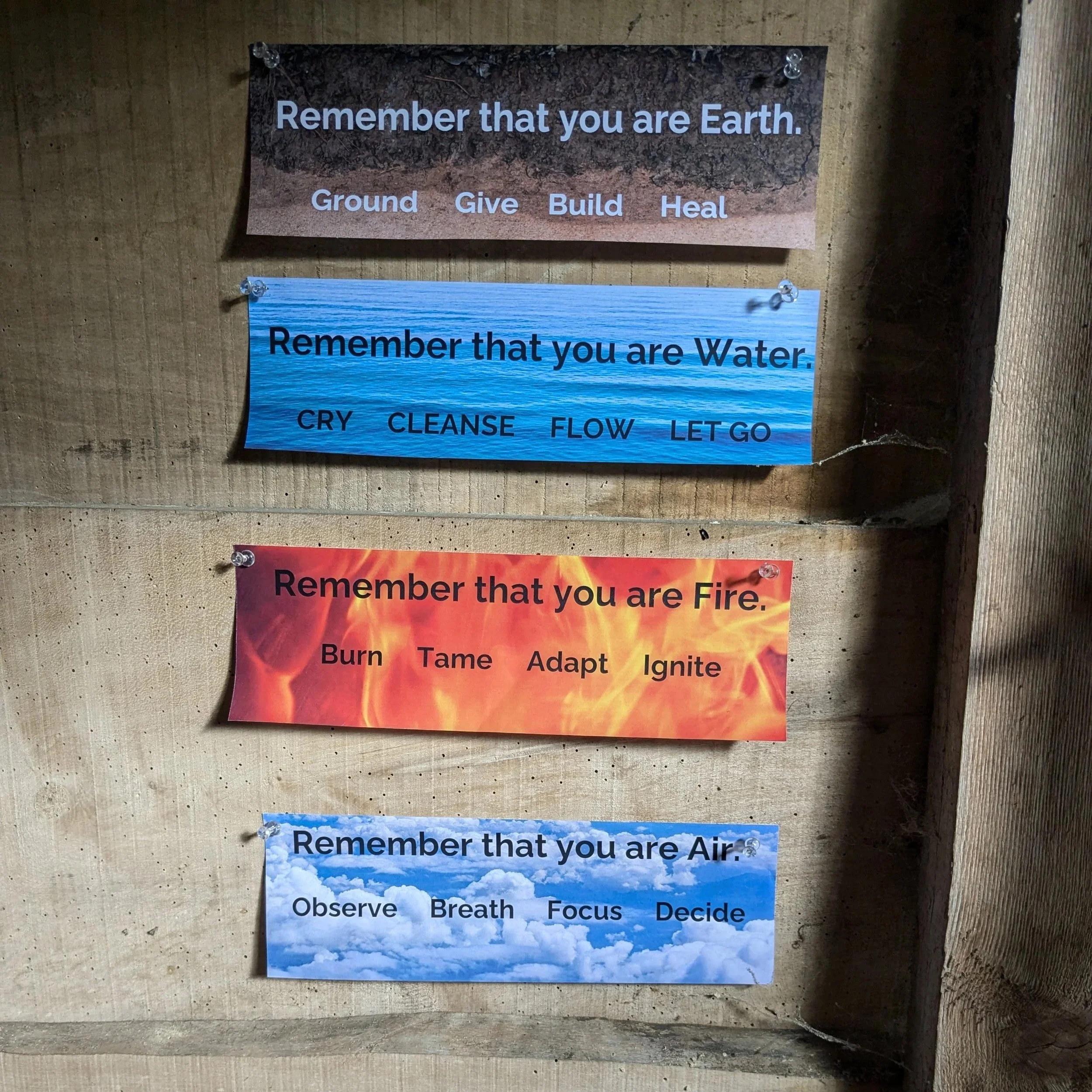 A series of signs with nature-themed backgrounds, each conveying a reminder: the top sign says "Remember that you are Earth" with soil imagery and words 'Ground,' 'Give,' 'Build,' 'Heal'; the middle sign says "Remember that you are Water" with water imagery and words 'Cry,' 'Cleanse,' 'Flow,' 'Let go'; the bottom sign says "Remember that you are Air" with sky imagery and words 'Observe,' 'Breathe,' 'Focus,' 'Decide'.