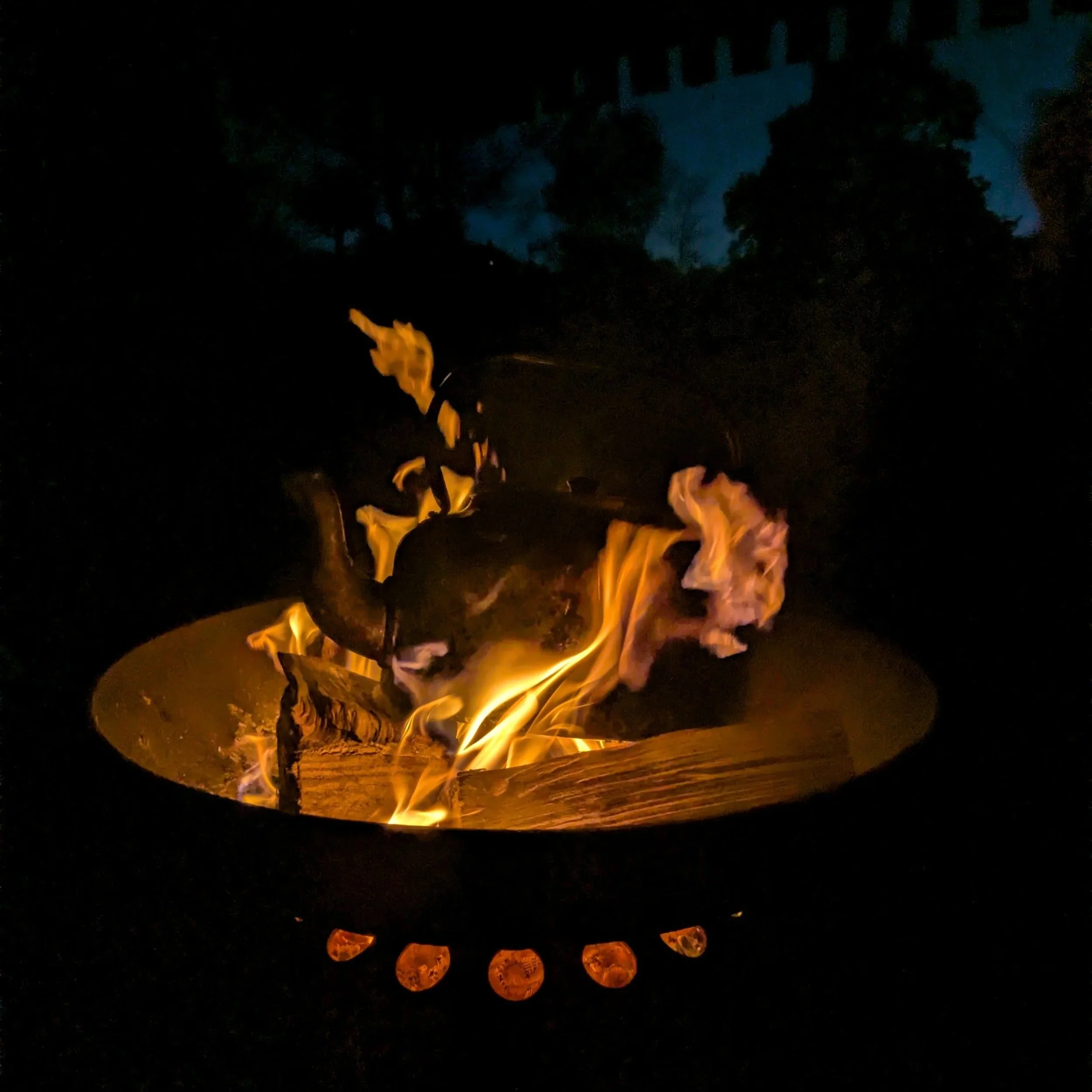 Nighttime scene with a fire burning and glowing orange flames, surrounded by darkness and silhouetted trees or structures in the background.