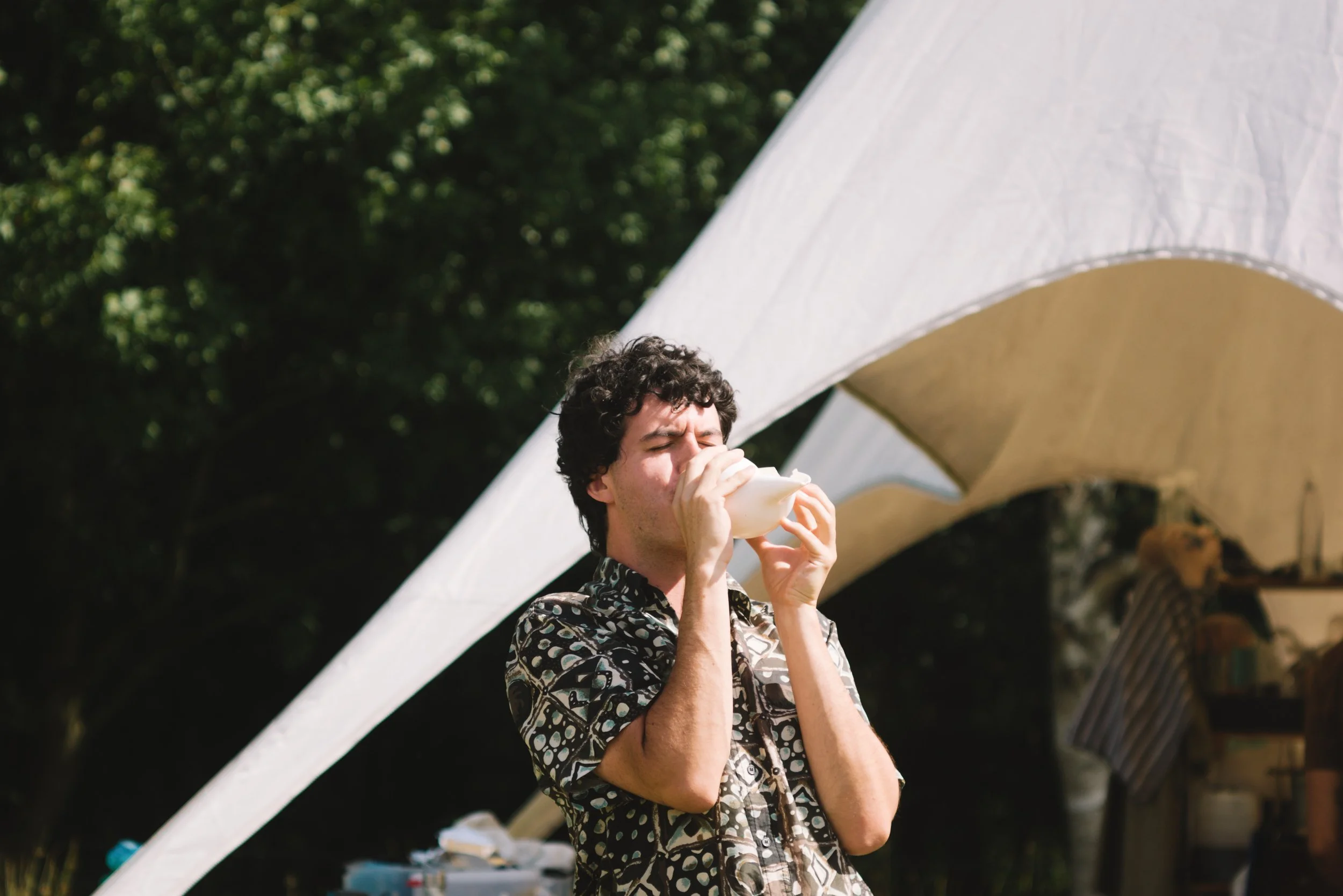 A young man with curly dark hair drinking from a white cup outdoors during daytime. He is wearing a patterned shirt, and there is a large white tent behind him. The background features green trees and a blurred outdoor setting.