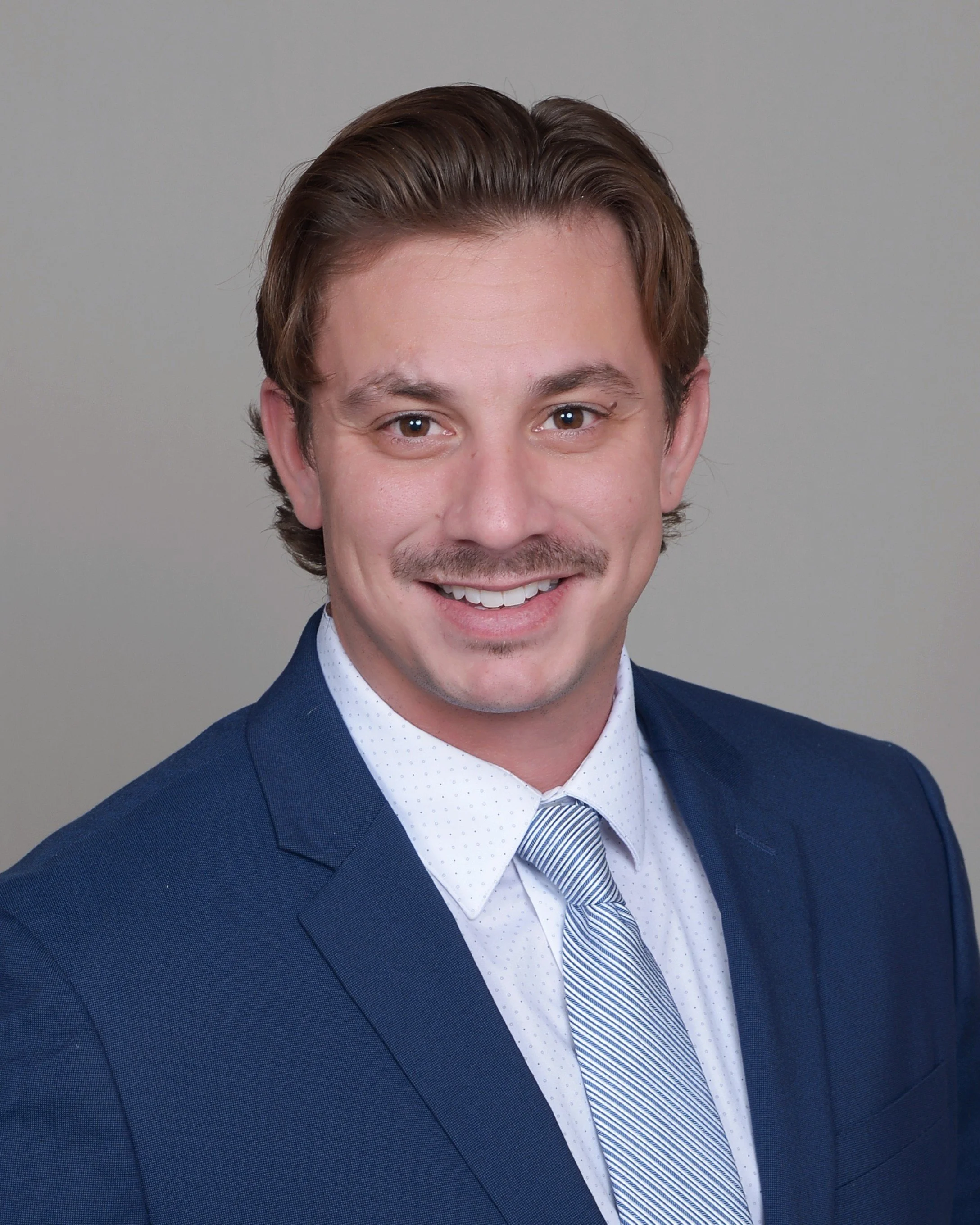 Close-up of AJ Gubash in a dark blue suit, white dress shirt, and striped tie, smiling against a plain gray background.