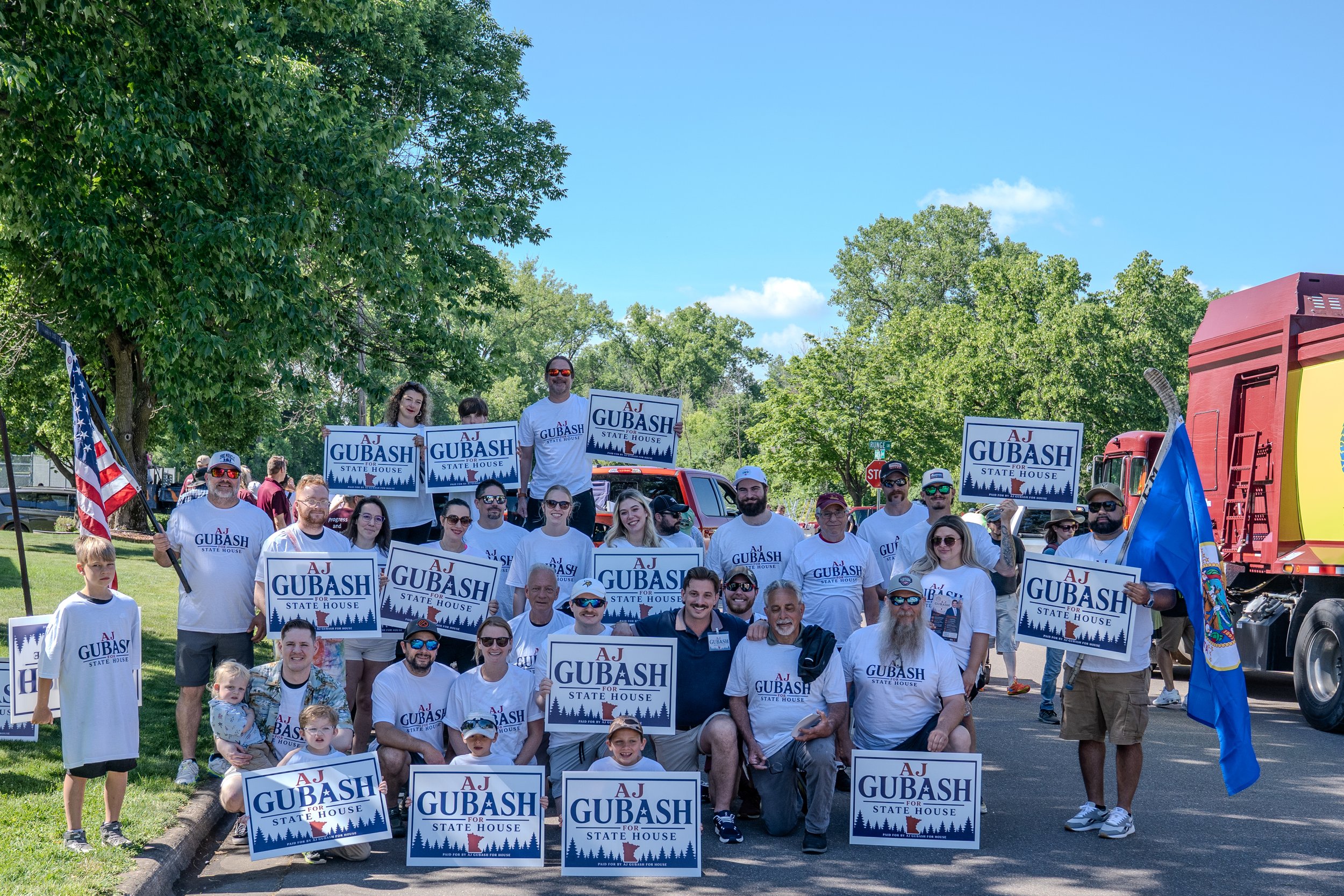 AJ Gubash and a group of people gathered outdoors holding campaign signs that say "AJ GUBASH for State House," with some standing on the back of a truck and others on the ground, in a park-like area with trees and blue sky.