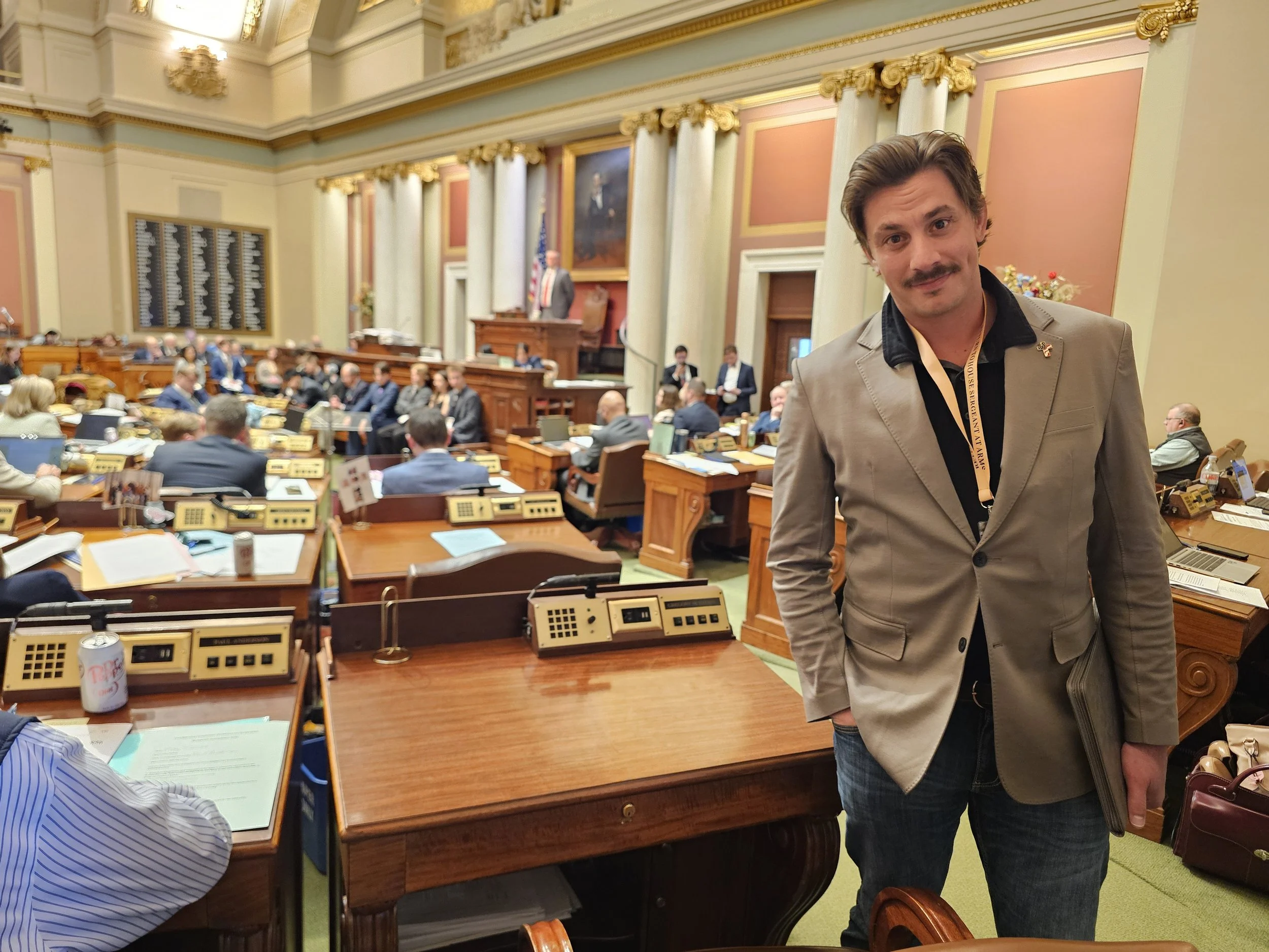 A man with a mustache and brown hair wearing a beige blazer and black shirt standing in the foreground of a large formal room with a legislative or parliamentary setting, filled with people seated at desks with computers and microphones, a large portrait on the wall, and a person standing at a podium in the background.