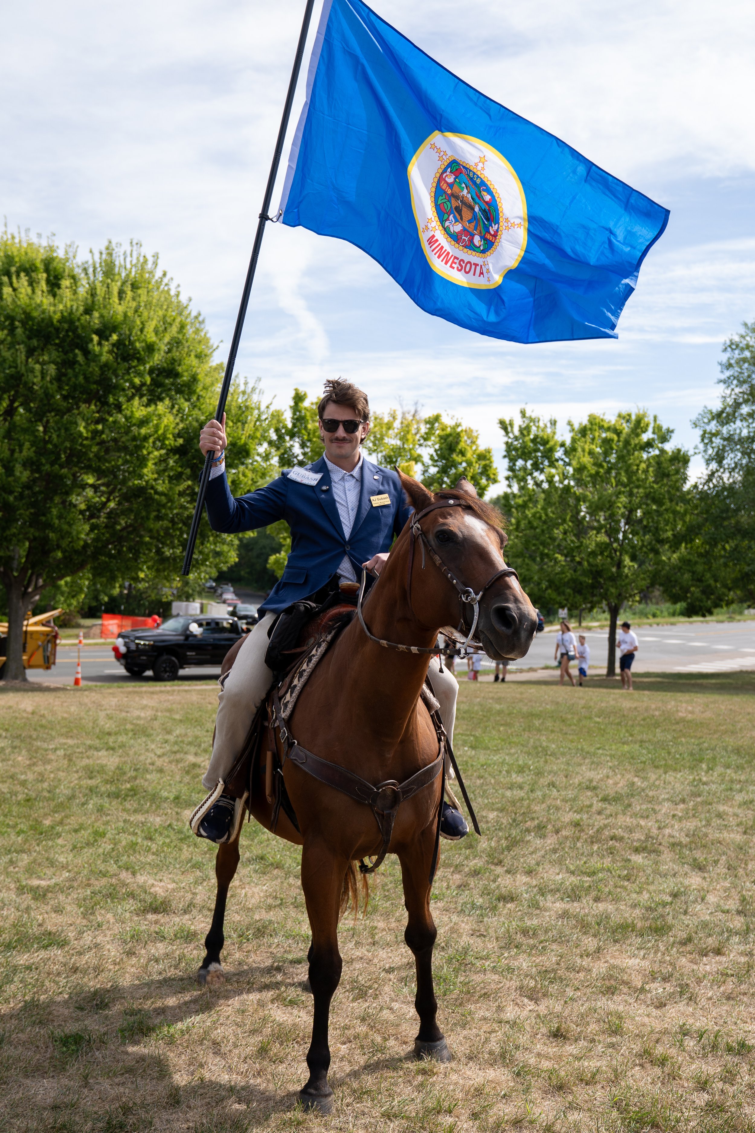 AJ Gubash wearing sunglasses and a blue suit riding a brown horse while holding a flag with the Minnesota state seal in an outdoor park area.