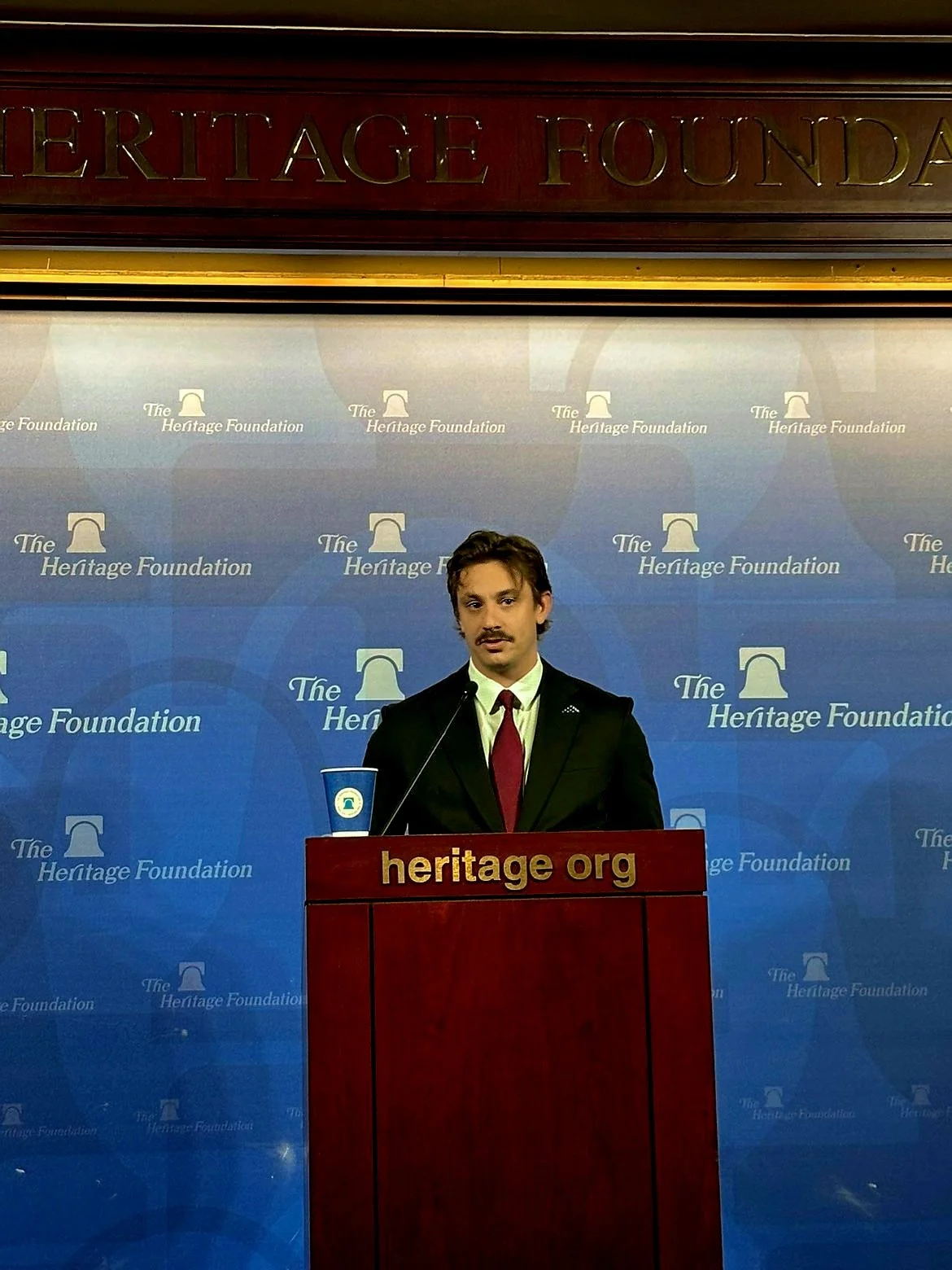 AJ Gubash in a black suit and red tie standing at a podium with a blue background that has 'The Heritage Foundation' logo. There is a sign on the podium that says 'heritage org' and a blue cup.