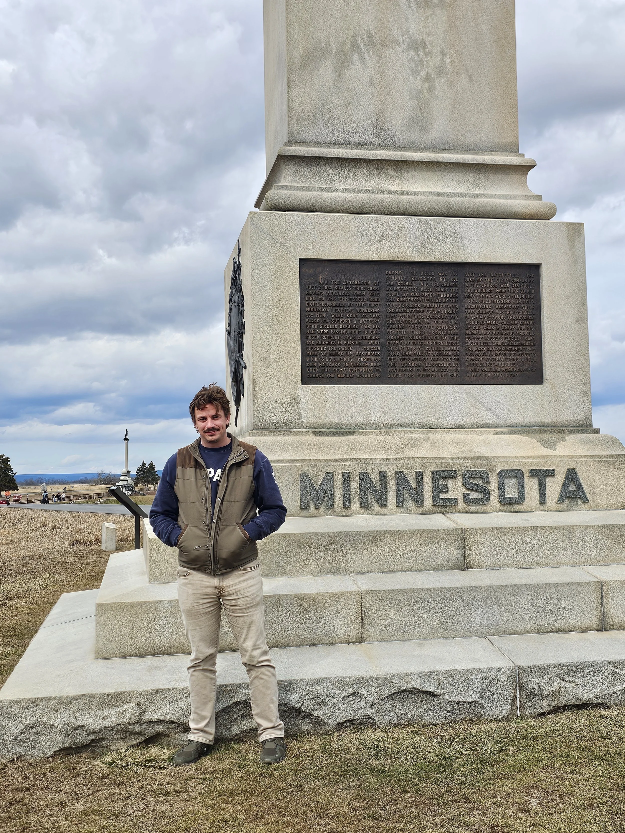 AJ Gubash standing in front of a large Minnesota statue with a plaque on it. The man is wearing a brown vest, dark blue hoodie, khaki pants, and black shoes. The background shows a cloudy sky, picnic tables, and a flag pole in a park setting.
