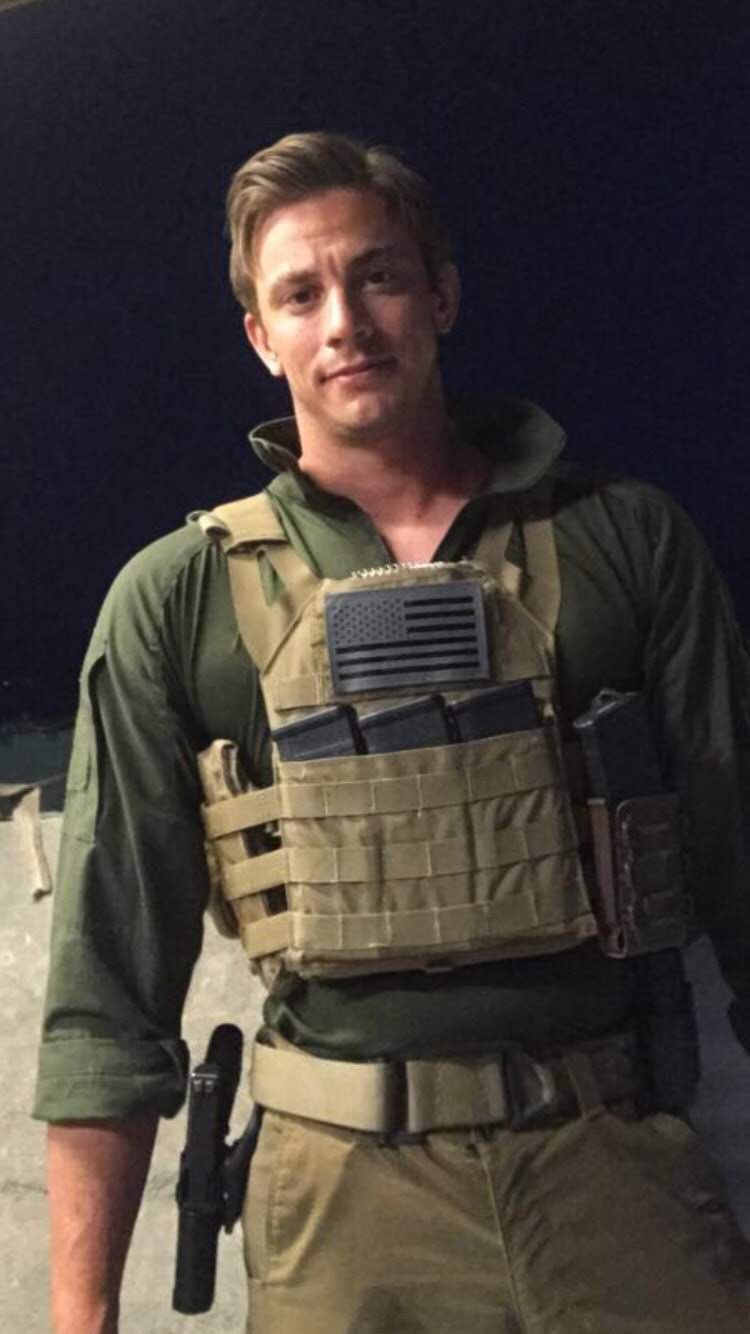 AJ Gubash in tactical military gear, including a bulletproof vest with magazines and a patch resembling the American flag, stands outdoors at night with a slight smile.