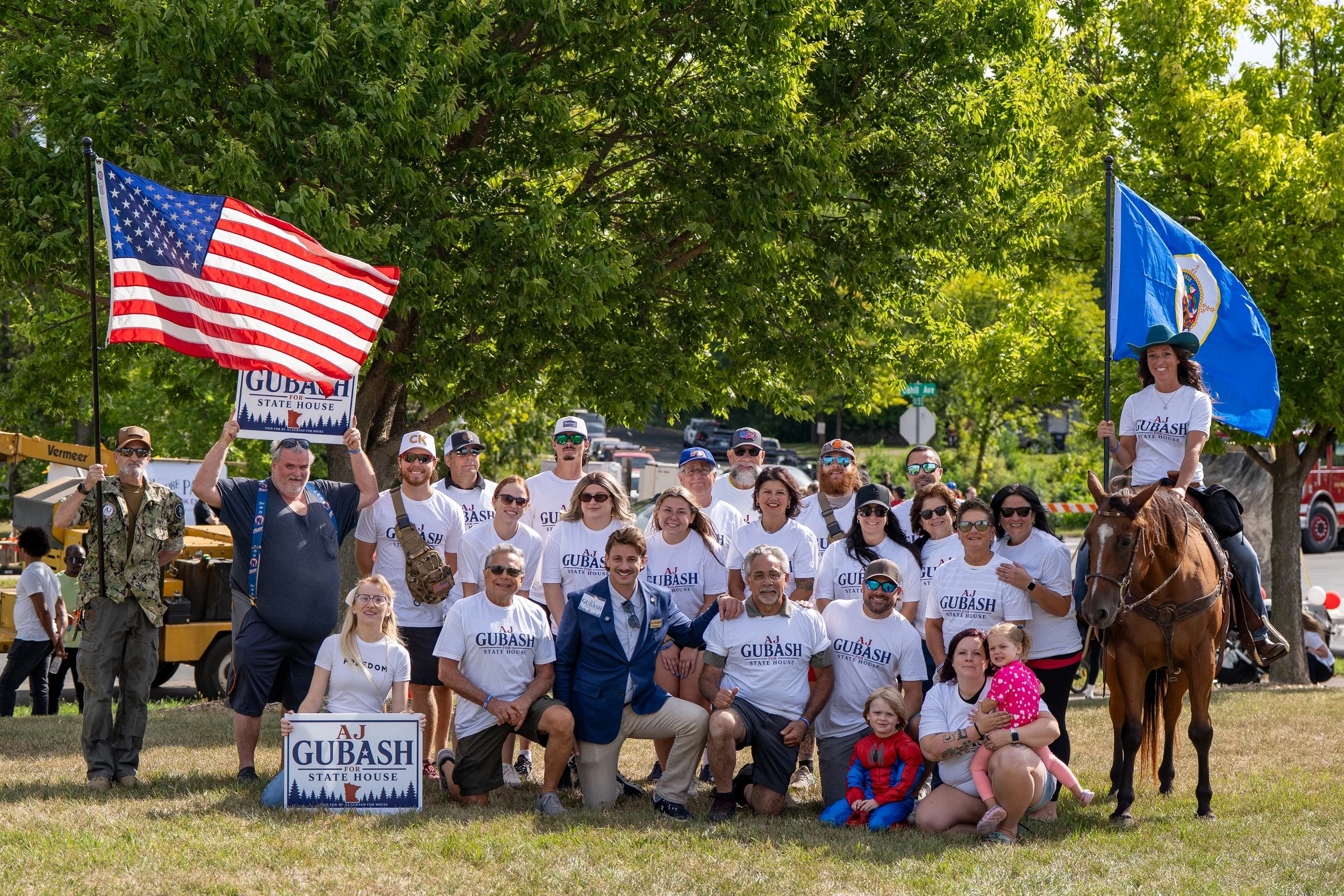 A large group of people gathered outdoors for a political rally, with some holding American flags and campaign signs supporting AJ Gubash for State House. The group includes people of various ages, some on horseback, and there are trees and a street in the background.