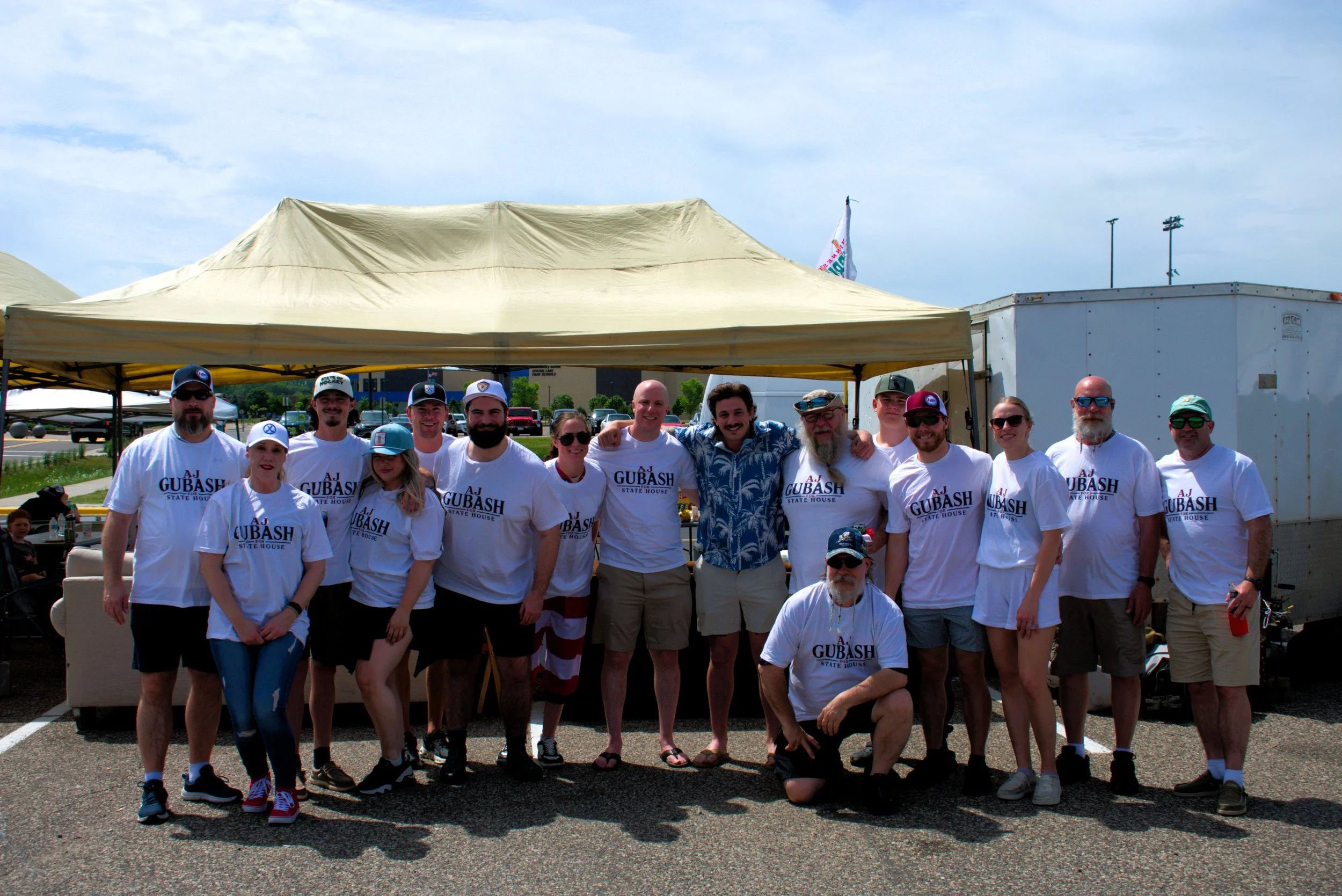 AJ Gubash and a group of people standing together outdoors under a tent, many wearing white t-shirts with 'AJ GUBASH STATE HOUSE' printed on them, posing for a photo at a public event.