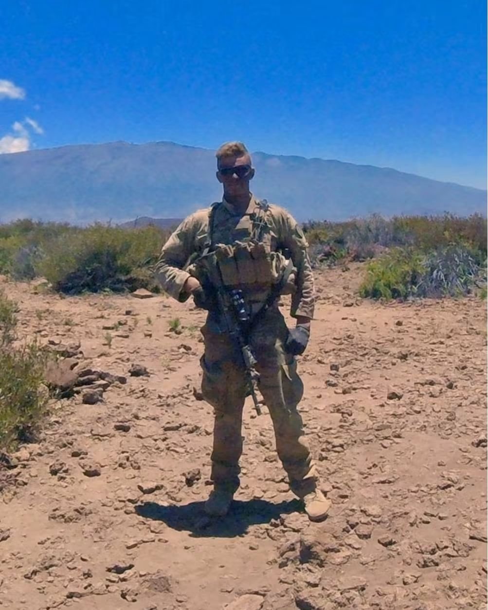 AJ Gubash wearing U.S. Army combat gear and sunglasses in a desert landscape with mountains in the background.