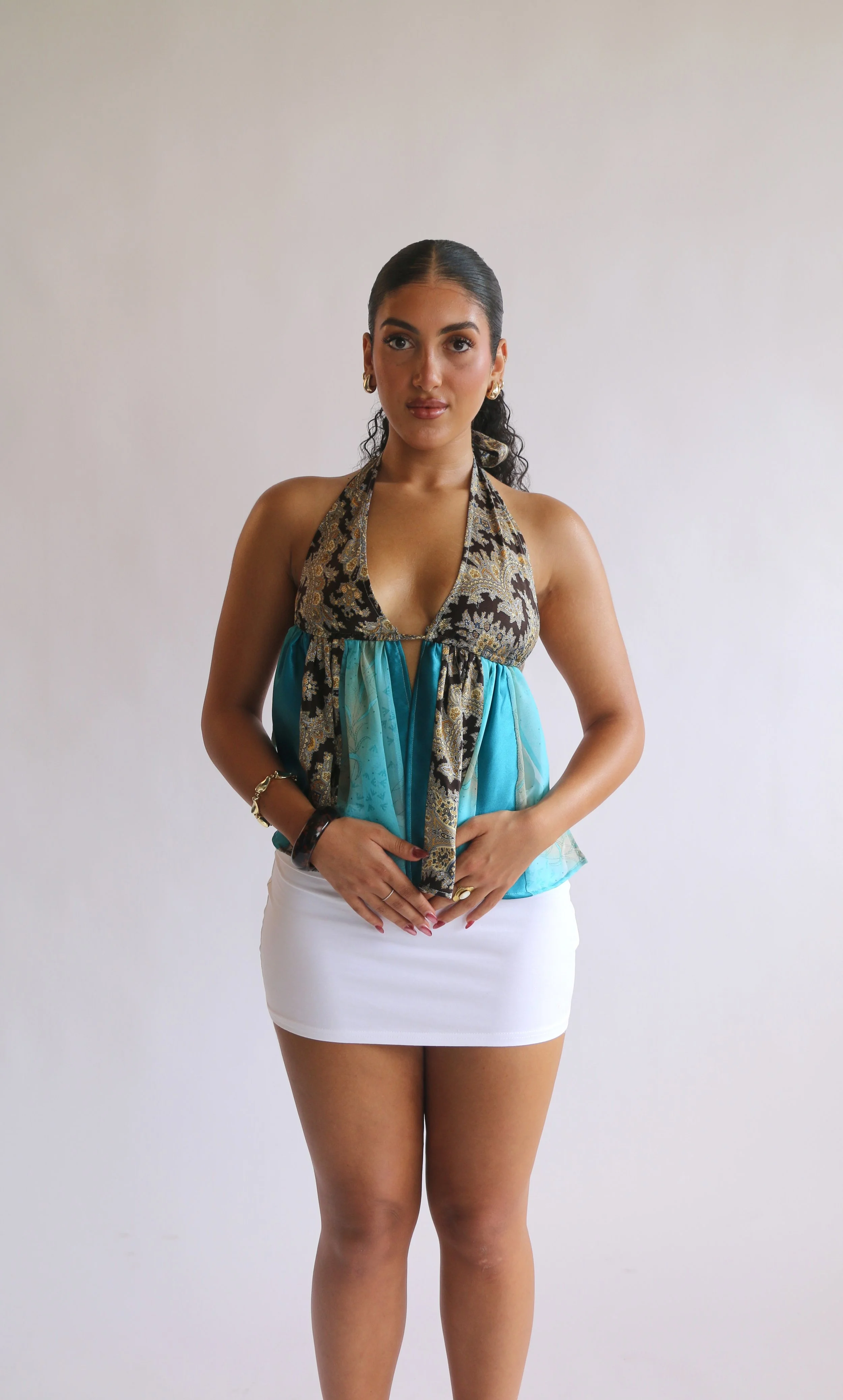 A woman standing against a plain light background, wearing a colorful halter top and a white skirt.