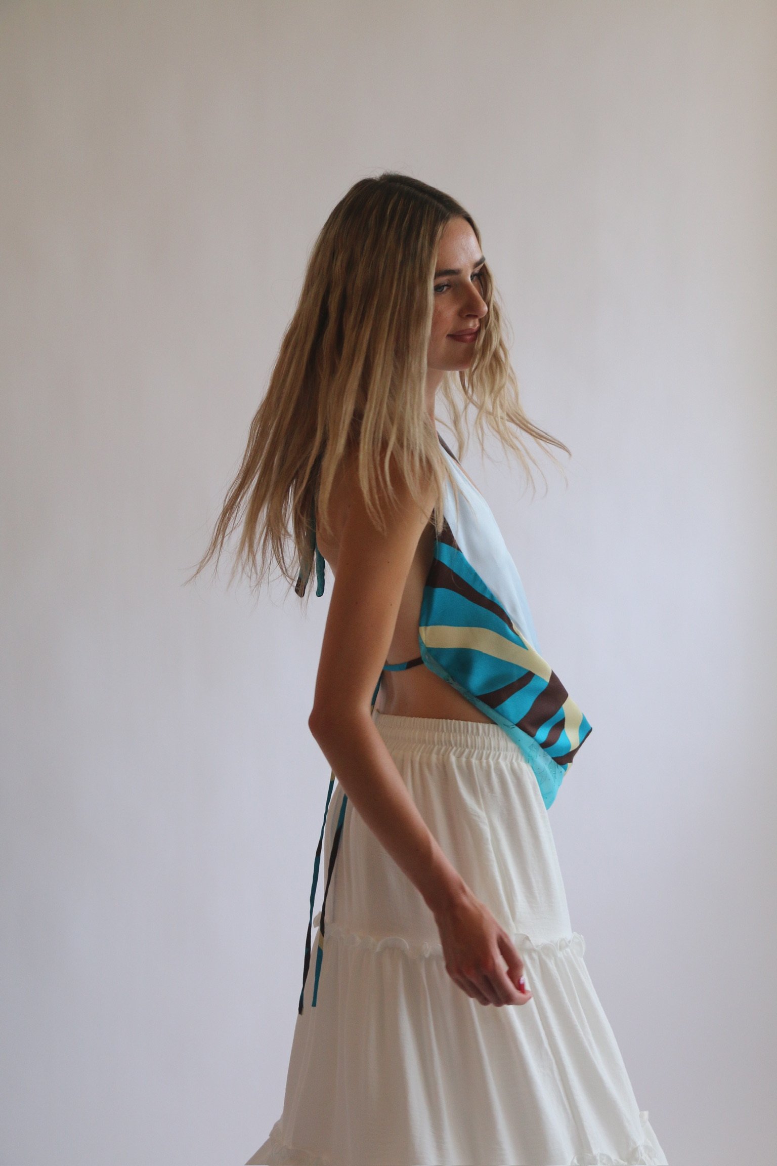 A young woman with shoulder-length wavy blonde hair, wearing a colorful striped top with shades of blue, brown, beige, and white, and cream-colored wide-leg pants, standing against a plain white wall.