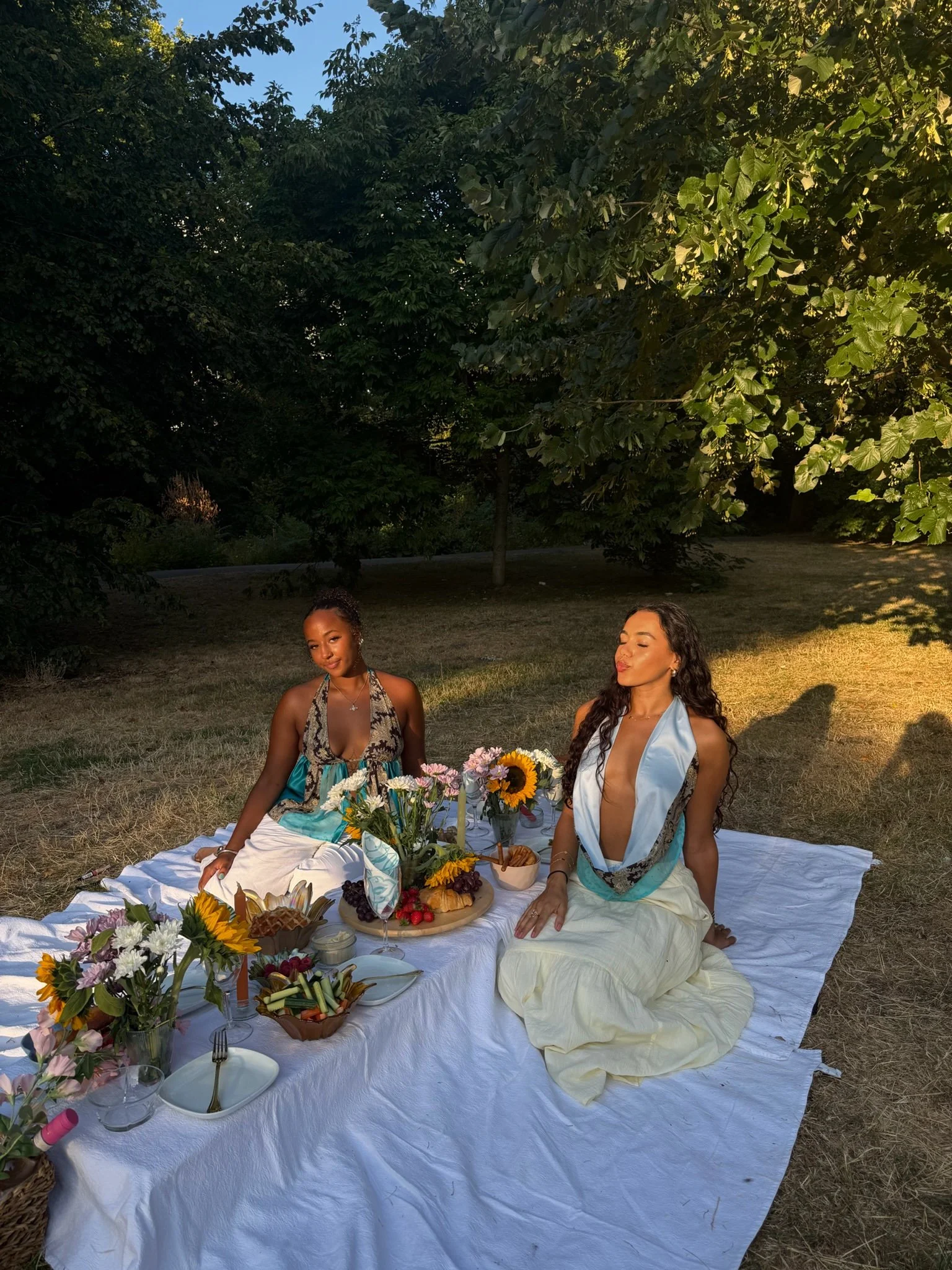 Two women sitting on a white blanket enjoying a picnic outdoors during the daytime, with a table filled with flowers, fruits, and food, surrounded by lush green trees.