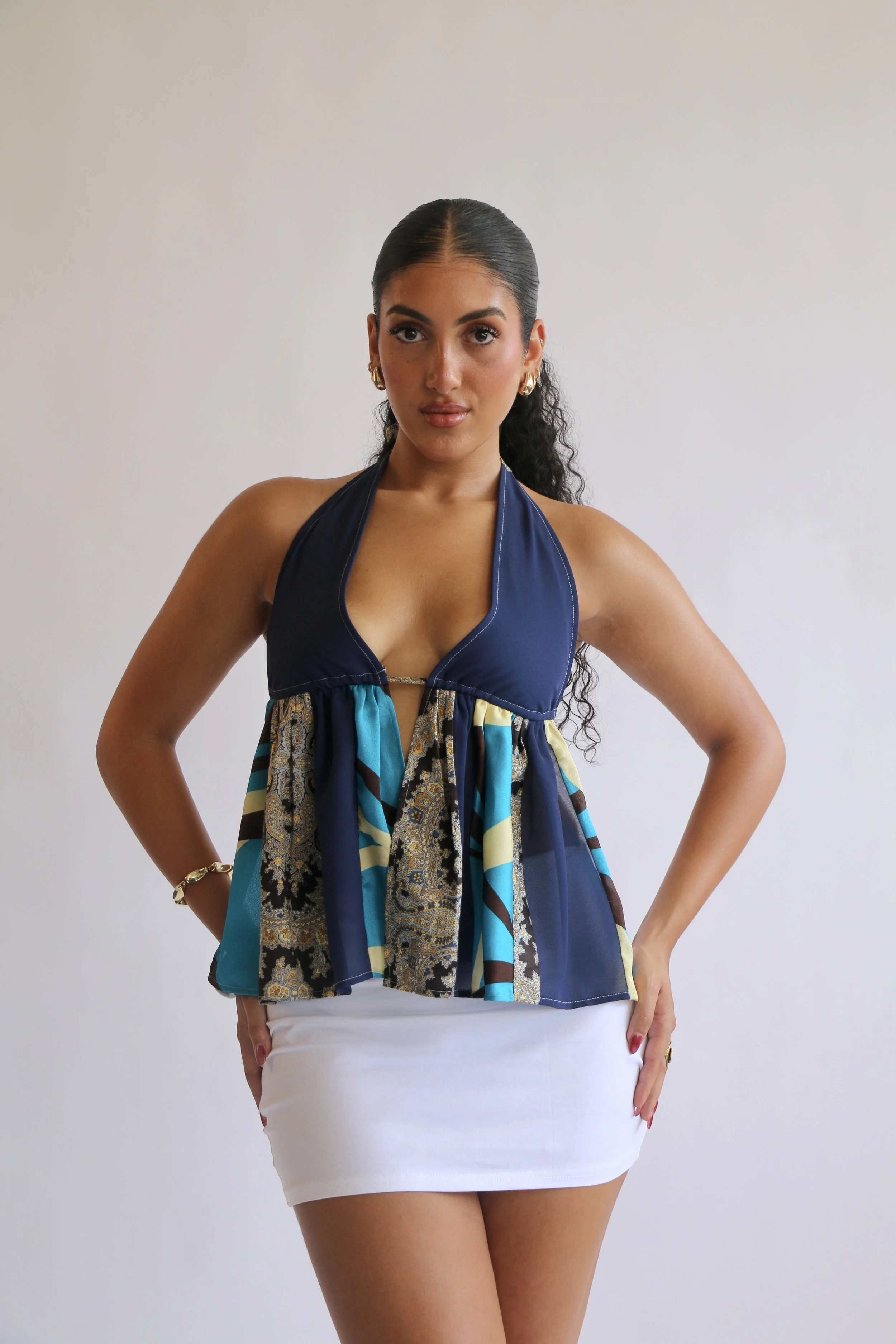 A young woman with dark, curly hair tied back, wearing a halter top with a multicolored pattern and gold earrings, standing with hands on hips against a plain white wall.