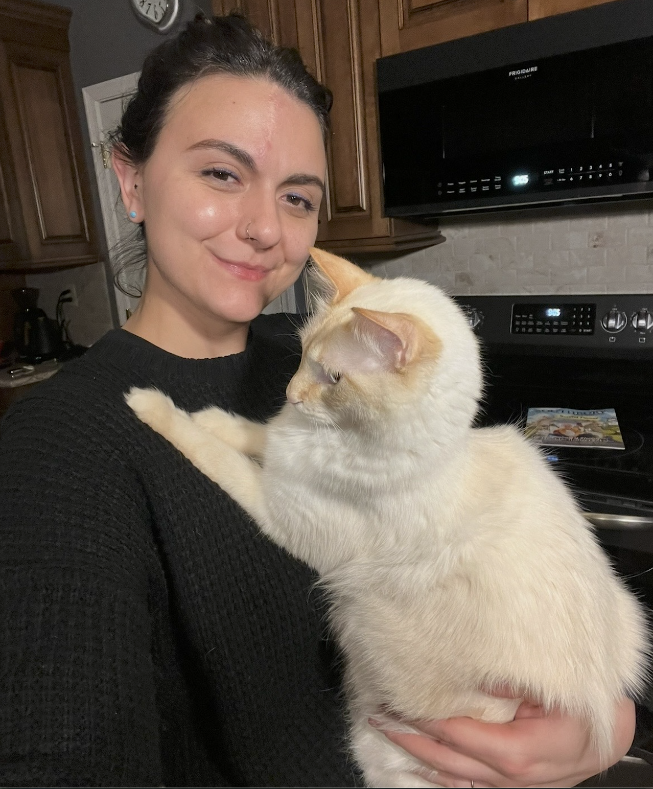 A woman smiling while holding a white and orange cat in a kitchen. The woman has dark hair, blue earrings, and wears a black sweater. The kitchen has wooden cabinets, a digital clock, and a magazine on the stove.