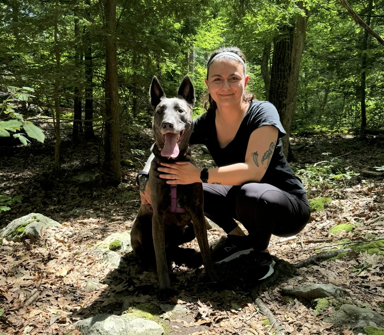 A woman and a dog in a forest. The woman is crouching and holding the dog, both smiling. The woman is wearing a black T-shirt, black pants, and a headband. The dog is a large, athletic breed with a tan and gray coat, and its tongue is out.