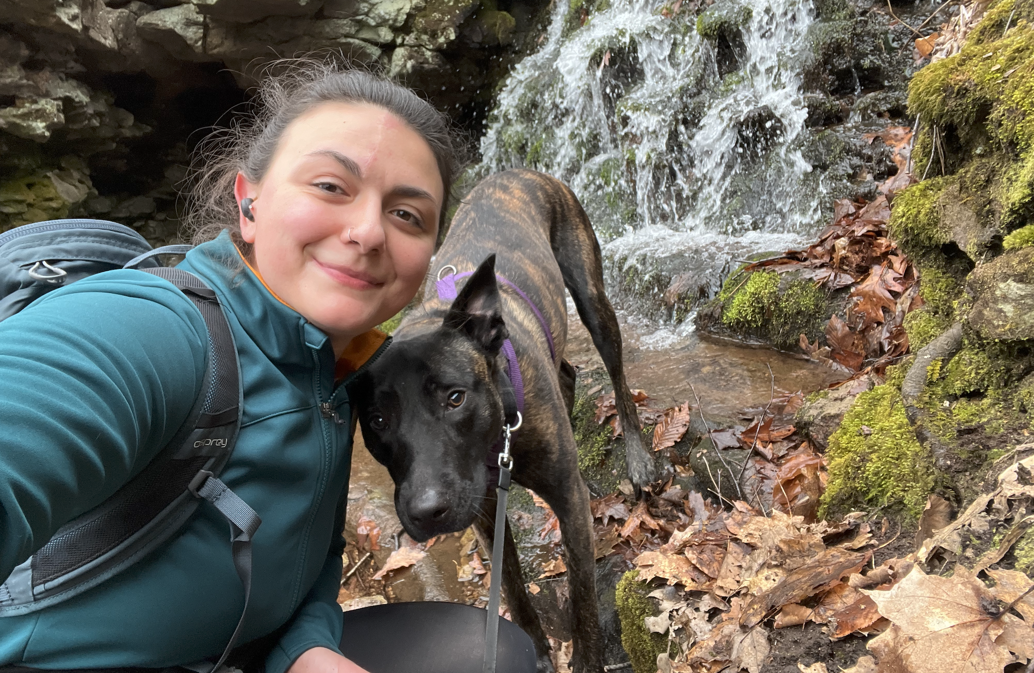 A young woman with a backpack taking a selfie with a black and brindle dog near a small waterfall surrounded by moss and fallen leaves in a forest.