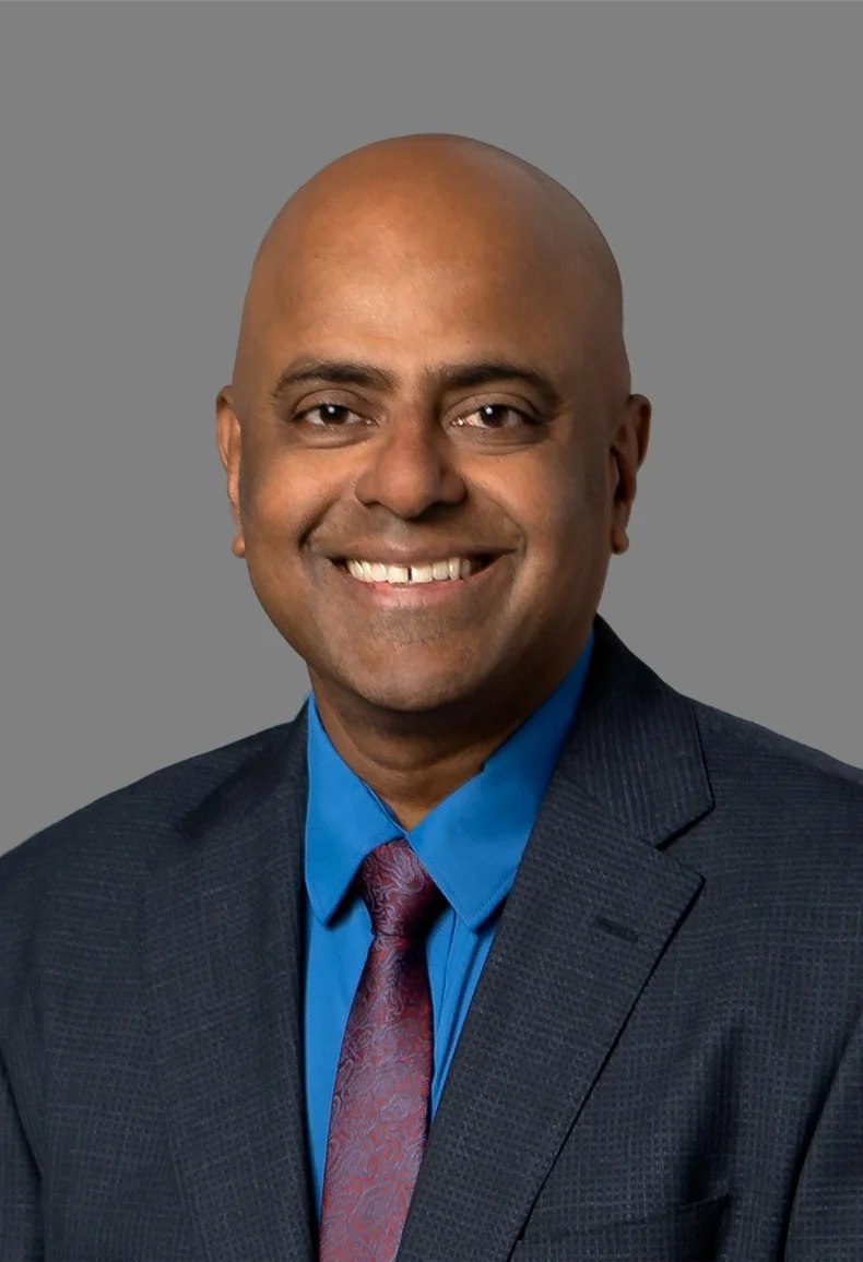 Headshot of a smiling man with a shaved head, wearing a dark suit jacket, blue shirt, and maroon tie, against a gray background.