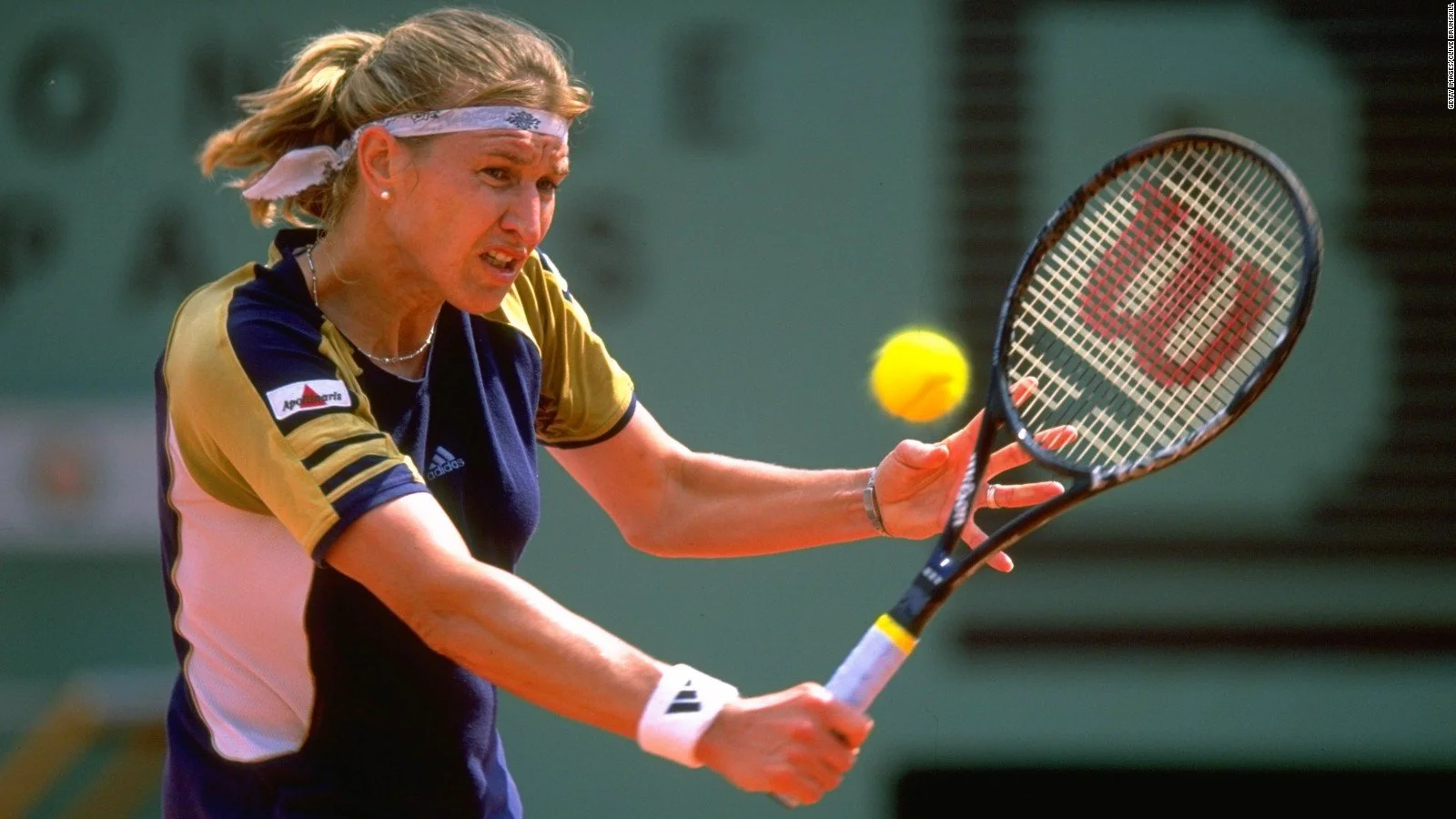 A woman with blonde hair, wearing a pink and navy blue sports shirt with an Adidas logo, is playing tennis and hitting a yellow ball with a tennis racket on a court.