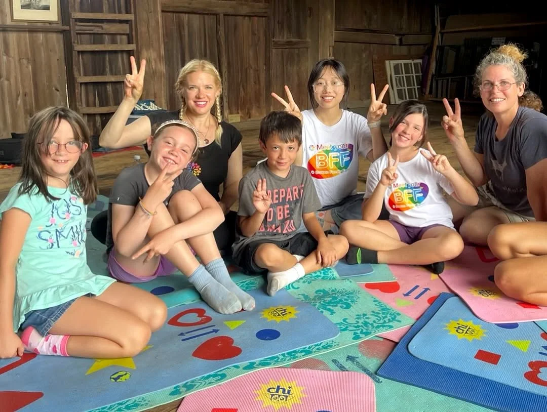 KaleidoscopeME ActiveLife Curriculum x Bowen Family Farm, Group of children and two adults sitting on colorful mats inside a wooden shed, smiling and making peace signs.