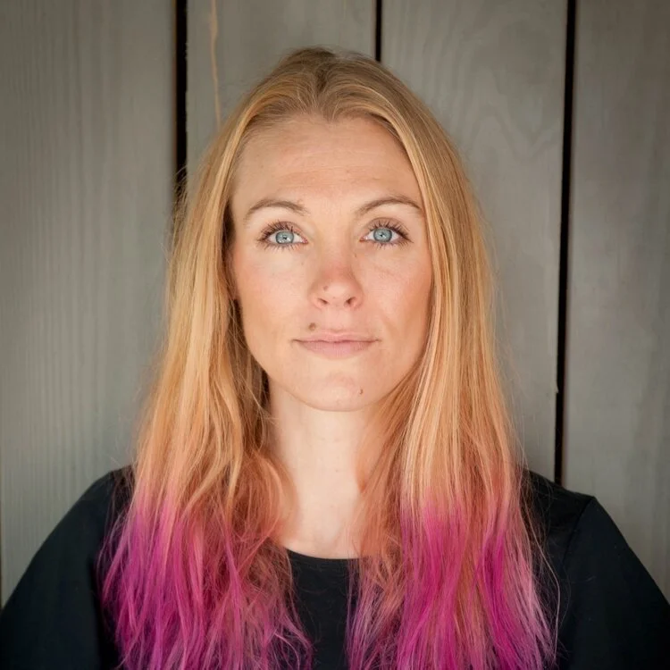 A young woman with long, wavy blonde hair fading into pink at the ends, wearing a black top, standing in front of a neutral wooden background.