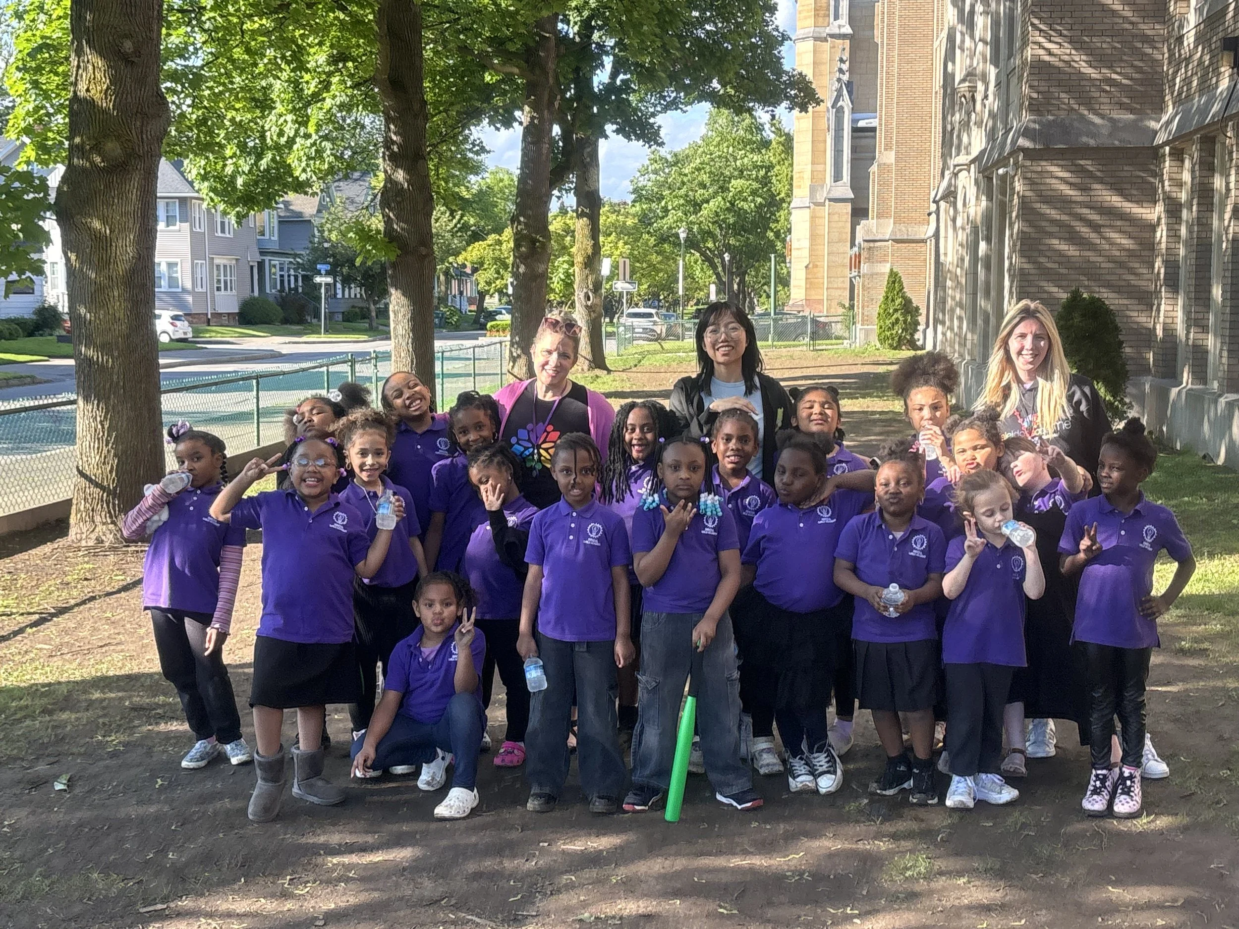 A group of children and two women standing outdoors on a sunny day in a park, with trees, a fence, and a brick building in the background. The children are wearing purple shirts, and some are holding water bottles. The women are smiling, one wearing glasses and the other with long blond hair.