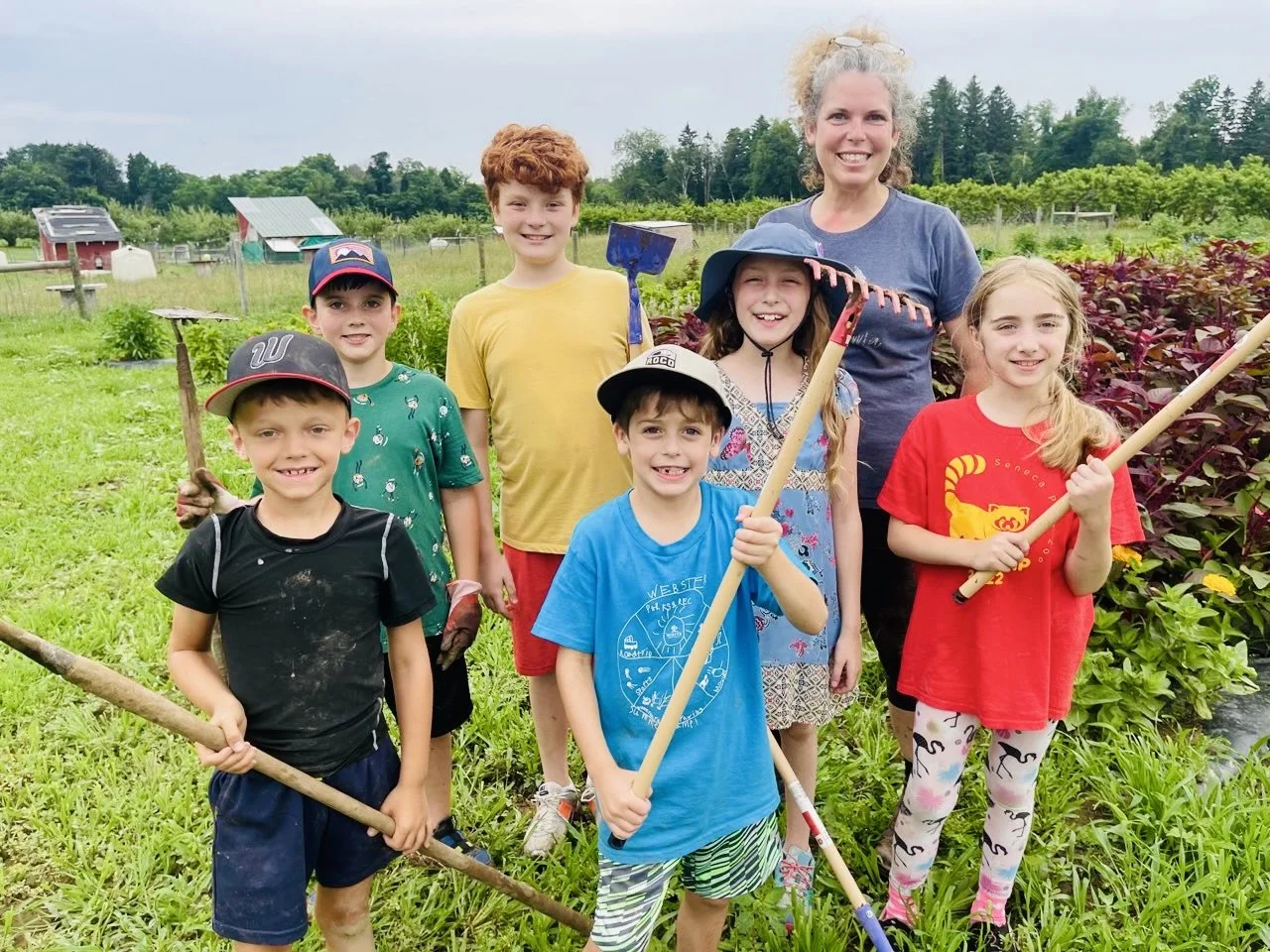 A group of six children and one adult woman standing in a garden, holding gardening tools like hoes and rakes, smiling at the camera.