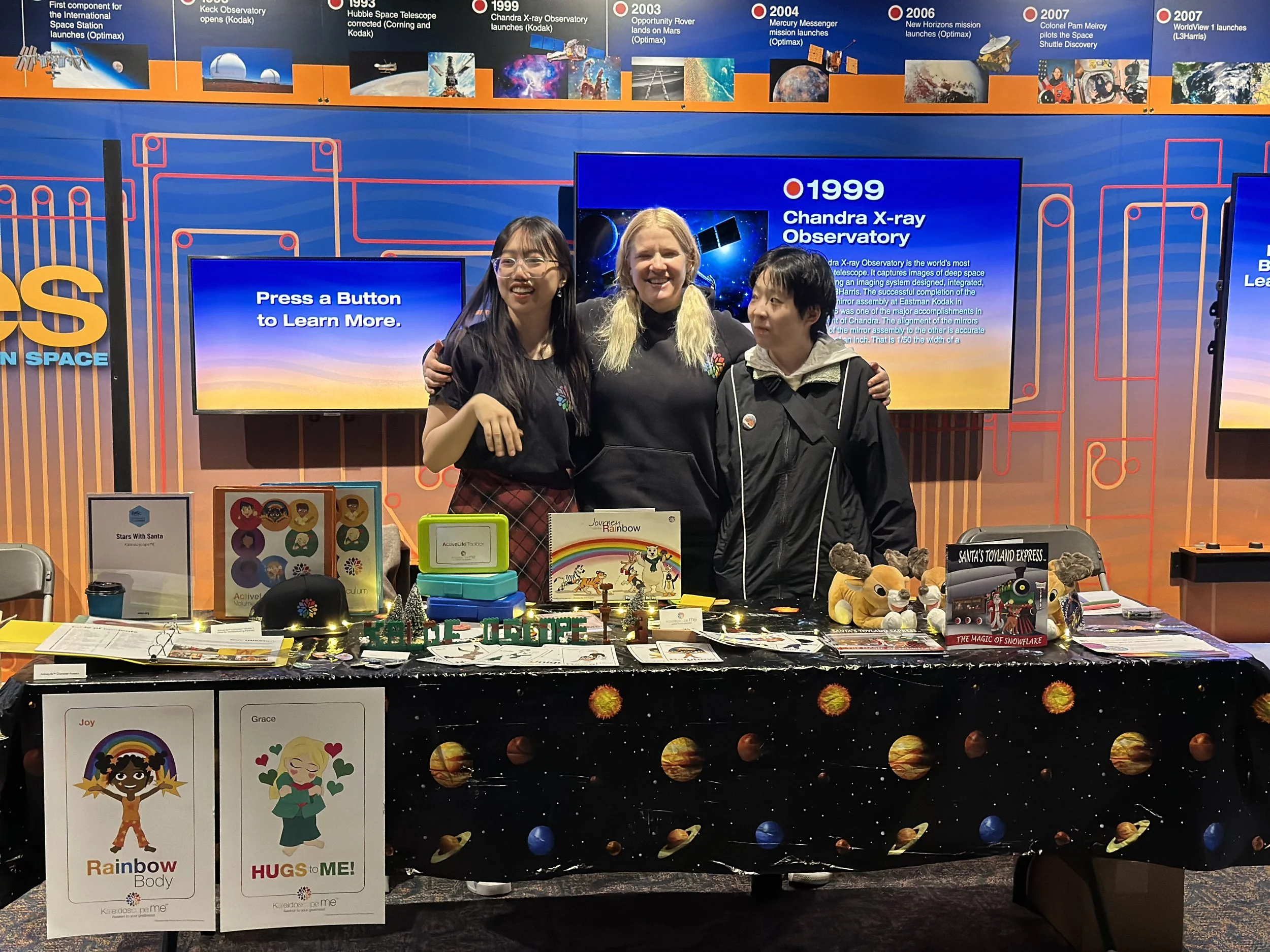 People standing behind a table with space-themed educational materials at a science exhibit. The background features a timeline of space exploration milestones, including images of spacecraft, telescopes, and planets, with various informational screens and posters.