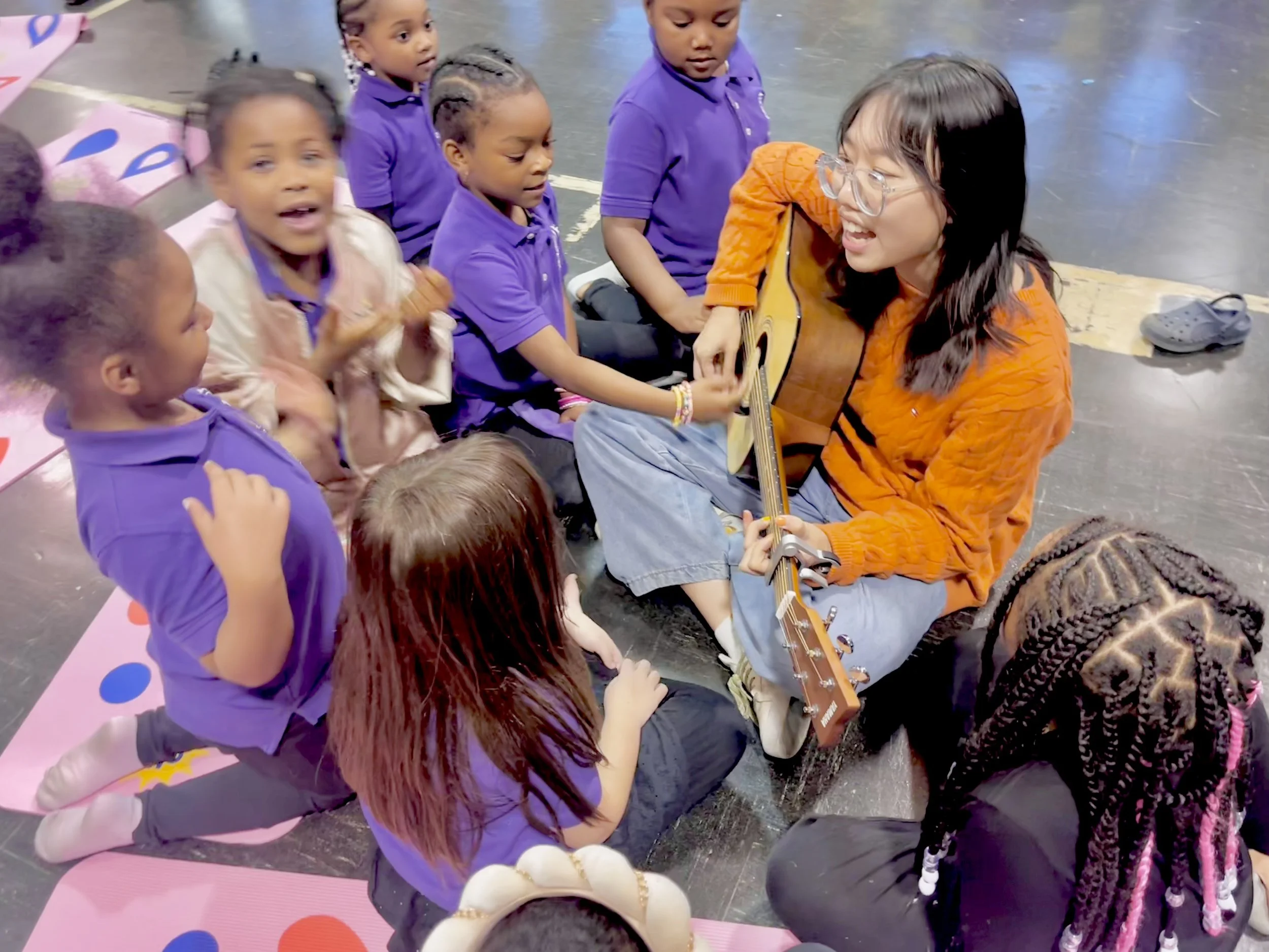 A group of young children sitting on the floor listening to a woman playing the guitar and singing. The children are wearing purple shirts and are sitting on a pink mat with colorful dots in a classroom or activity setting.