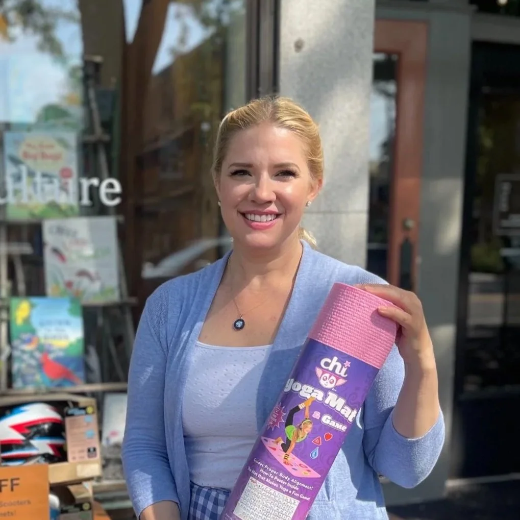 A woman with blonde hair, smiling and holding a pink yoga mat outside a store.