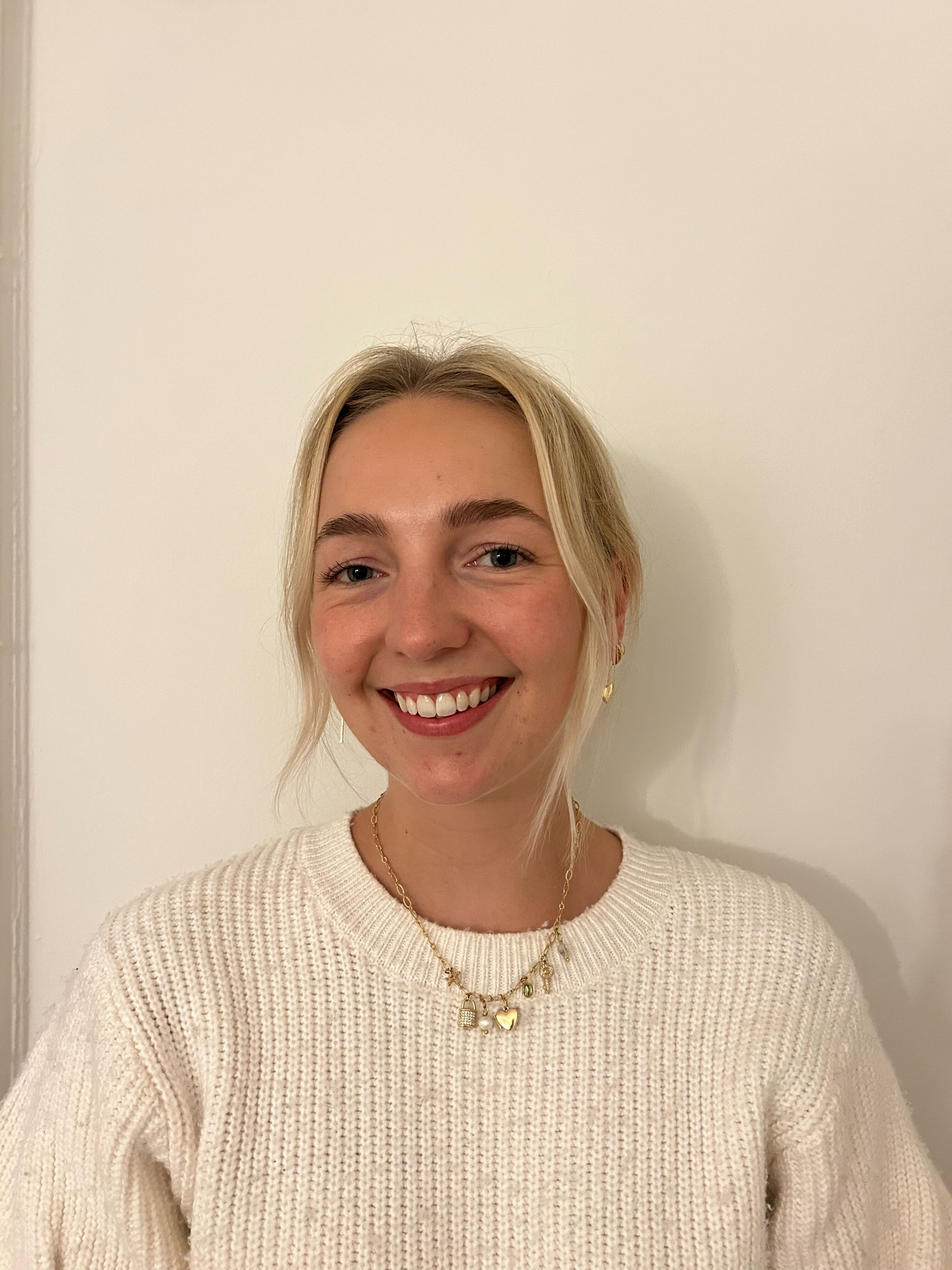 A smiling woman with blonde hair, blue eyes, wearing a cream sweater and layered necklaces, standing against a plain white wall.
