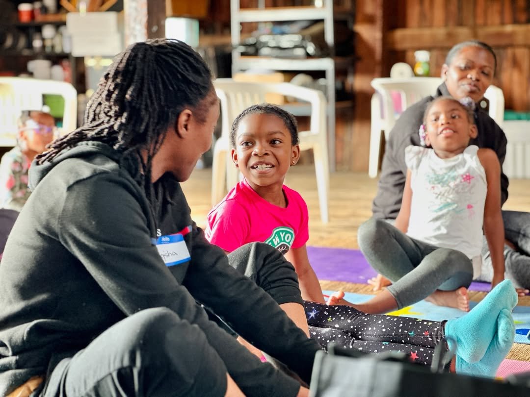 Three children and one adult sitting on the floor in a circle, engaging in conversation, with a background of shelves and chairs in a cozy indoor setting.