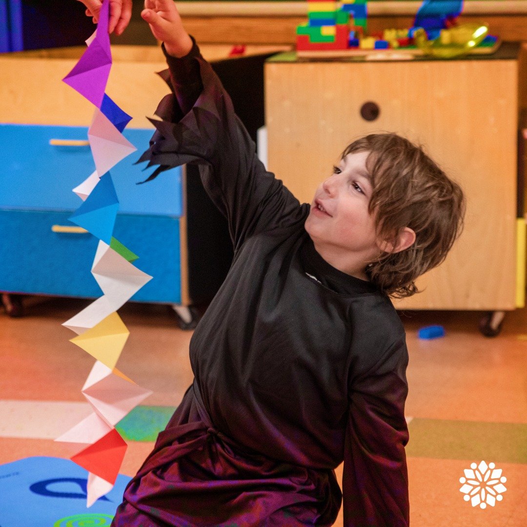 A young boy with brown, curly hair smiling while holding a colorful paper chain craft in a classroom setting with wooden cabinets and toys in the background.