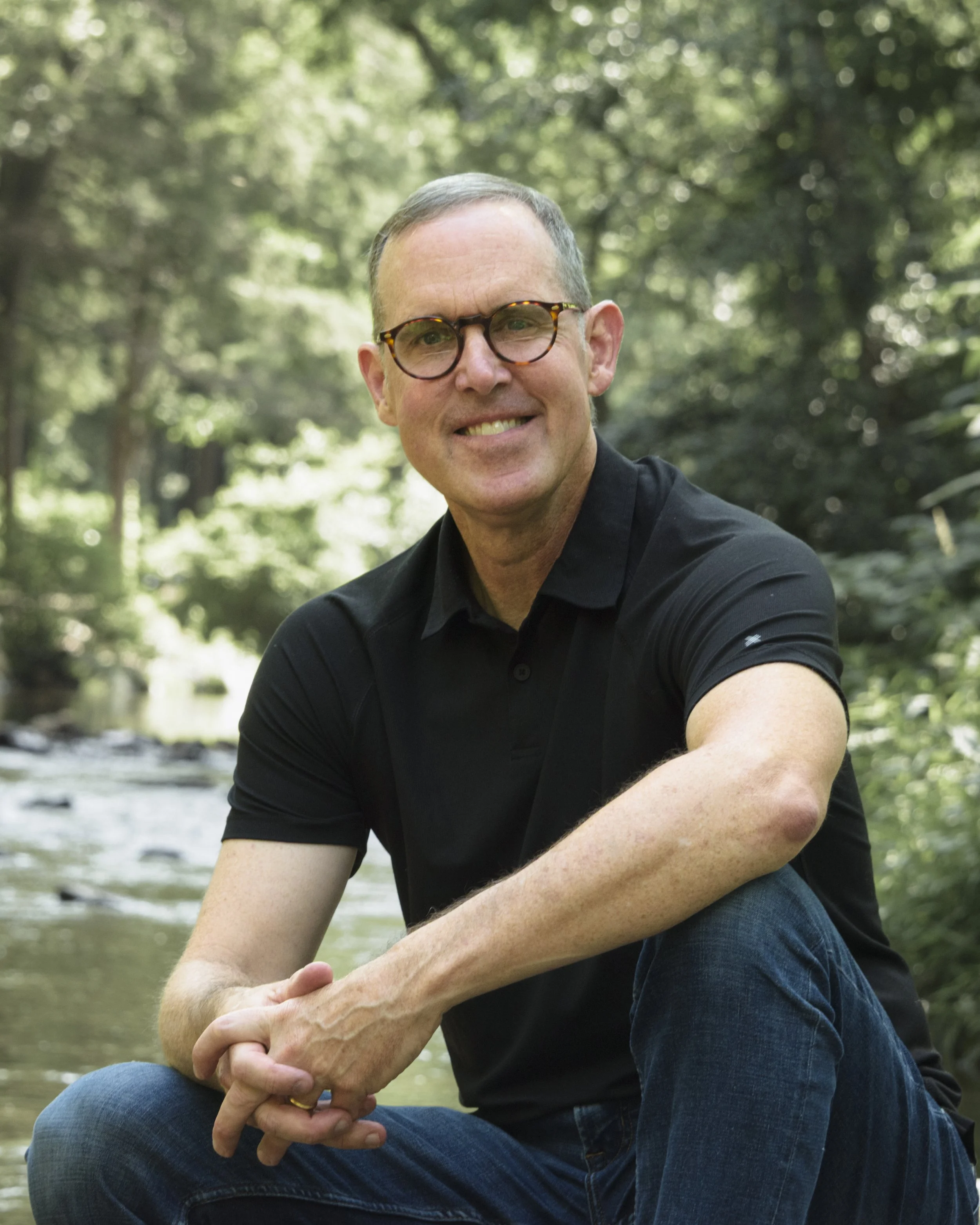 Smiling man wearing round glasses and a black polo shirt sitting beside a wooded stream