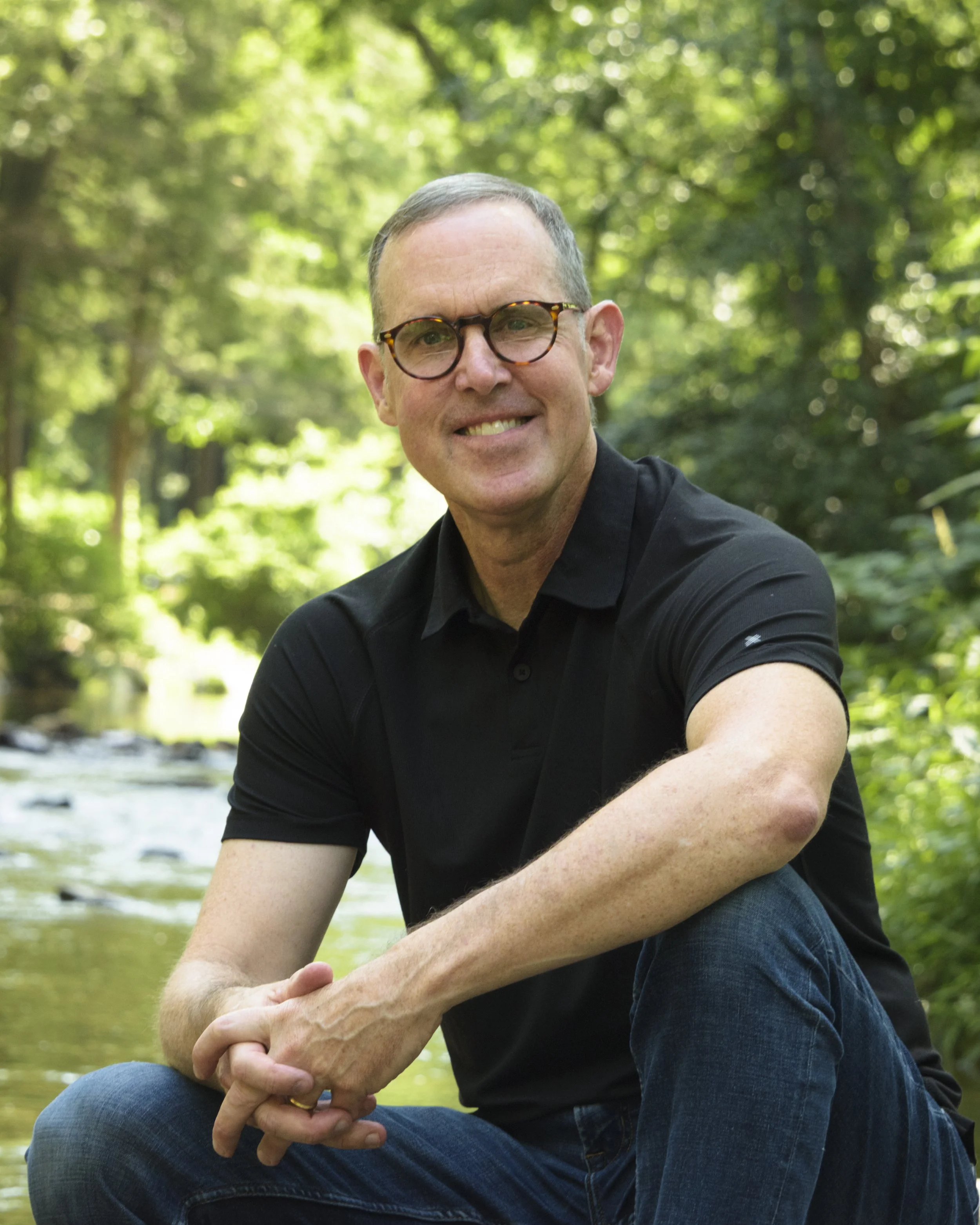 Smiling man wearing glasses and a black polo shirt sitting beside a wooded stream