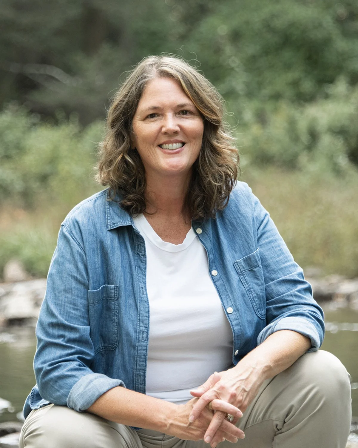 Smiling woman in a denim shirt sitting outdoors near a wooded stream