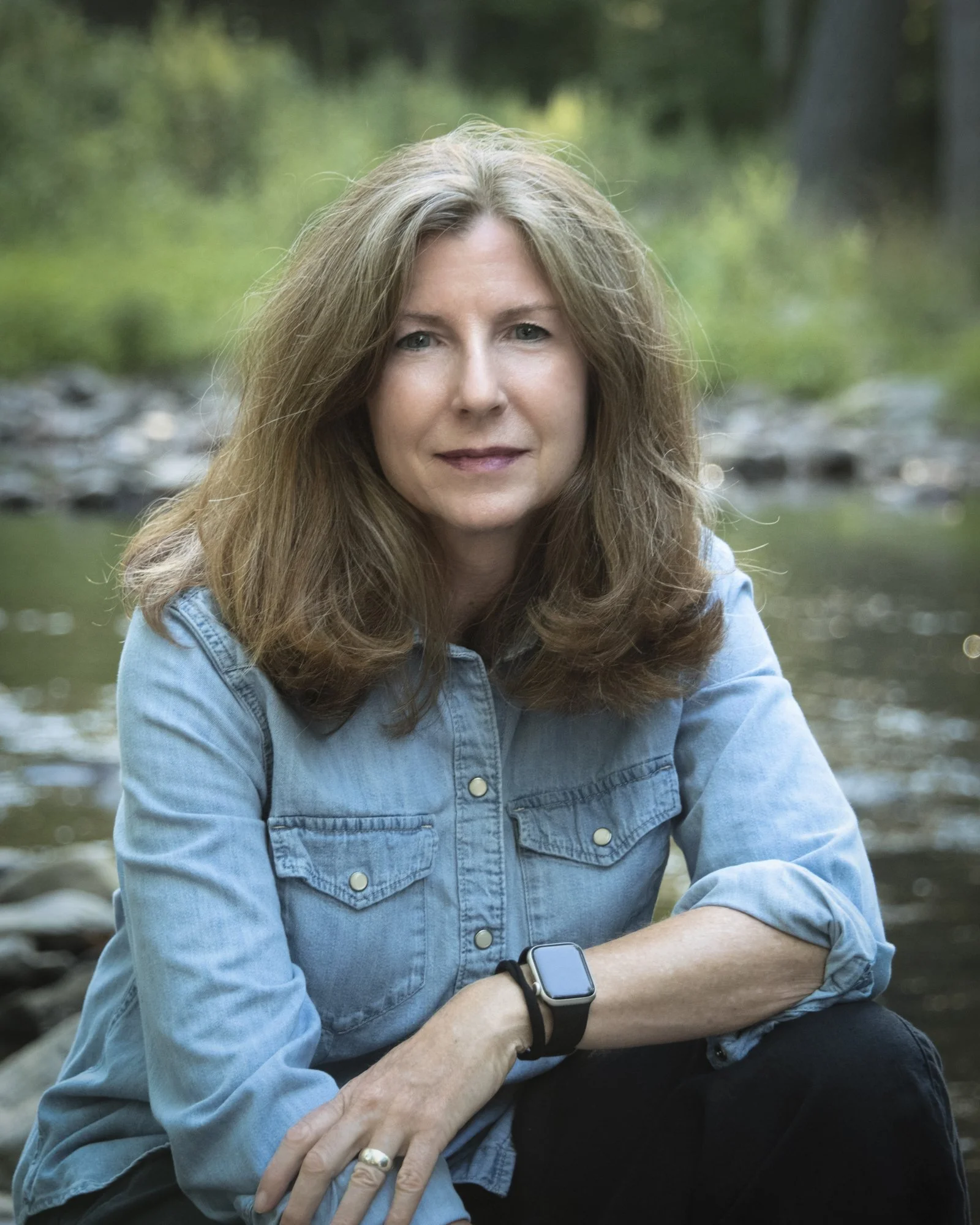 Portrait of a woman wearing a denim shirt beside a forest stream