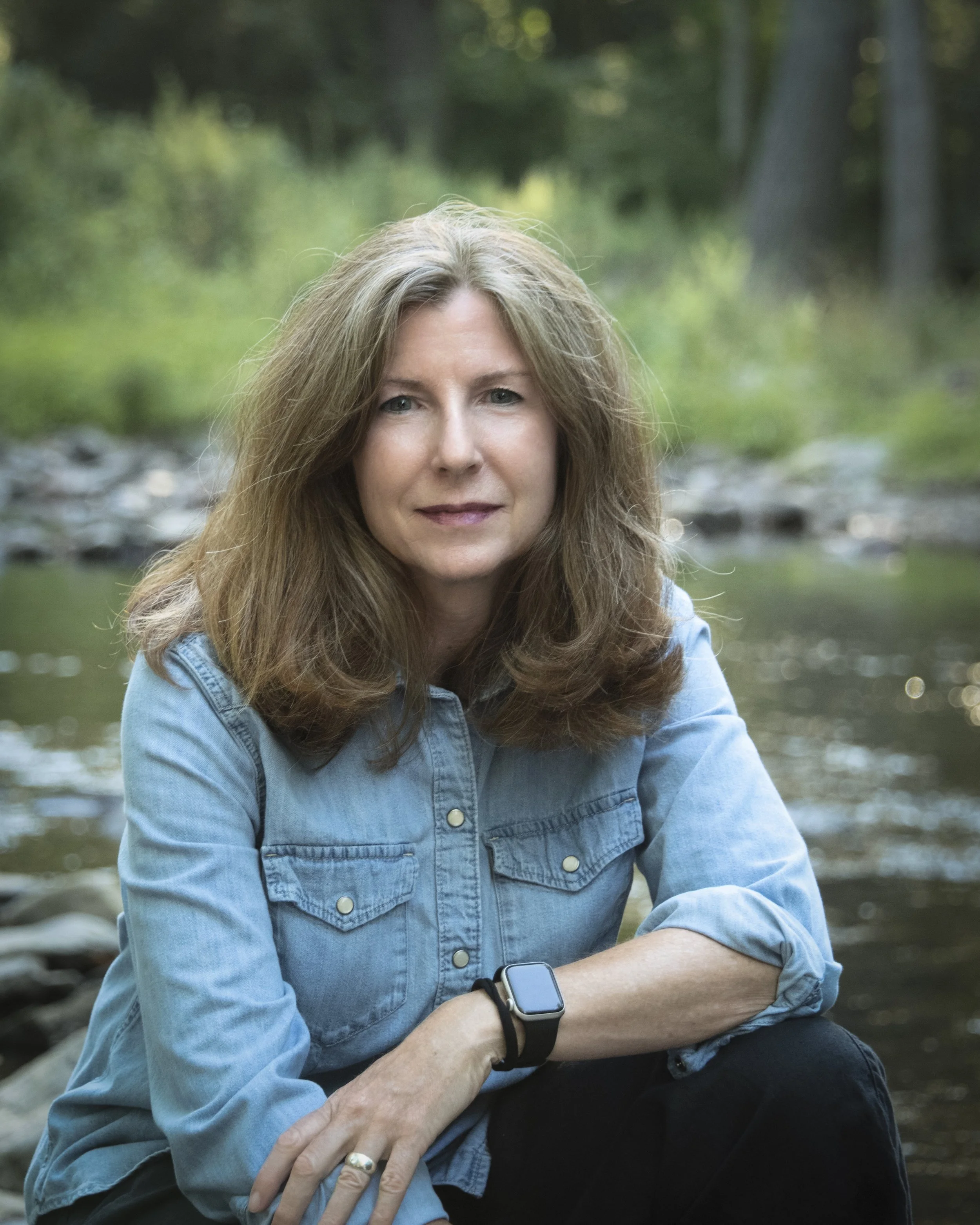 Woman in a denim shirt sitting outdoors beside a shallow stream
