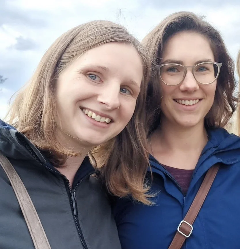 Zwei Frauen lächeln, während sie im Freien bei bewölktem Himmel stehen.