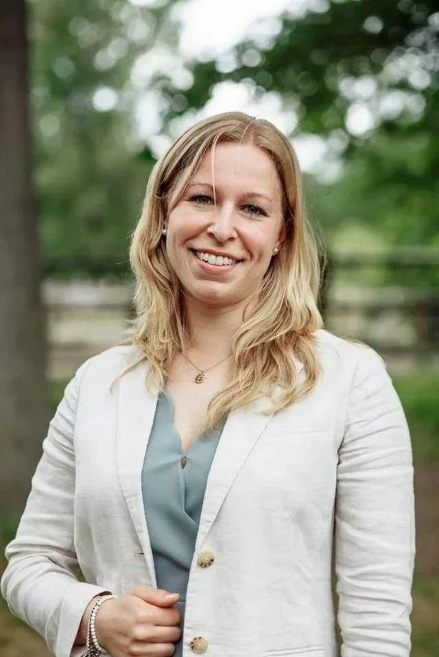 Eine Frau mit blondem Haar, die draußen vor einem Baum steht und in die Kamera lächelt.