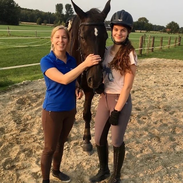 Zwei Mädchen und ein Pferd im Reitstall auf einer Sandpiste, eines der Mädchen trägt einen Reithelm.