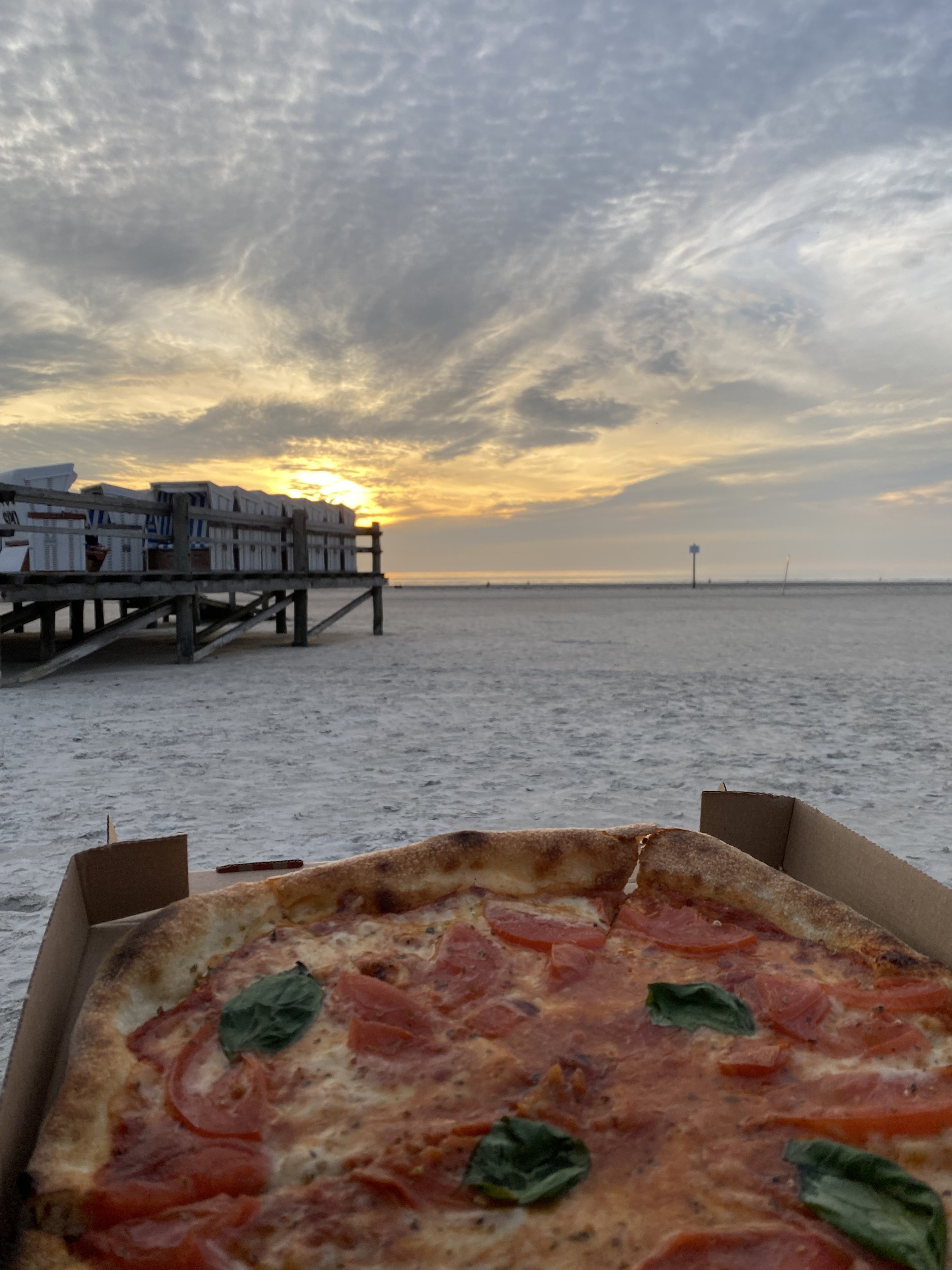Eine Pizza mit Tomaten, Basilikum und Käse in einer Pappschachtel auf einem Tisch am Strand bei Sonnenuntergang, im Hintergrund sind Strandliegen und der Strand sichtbar.