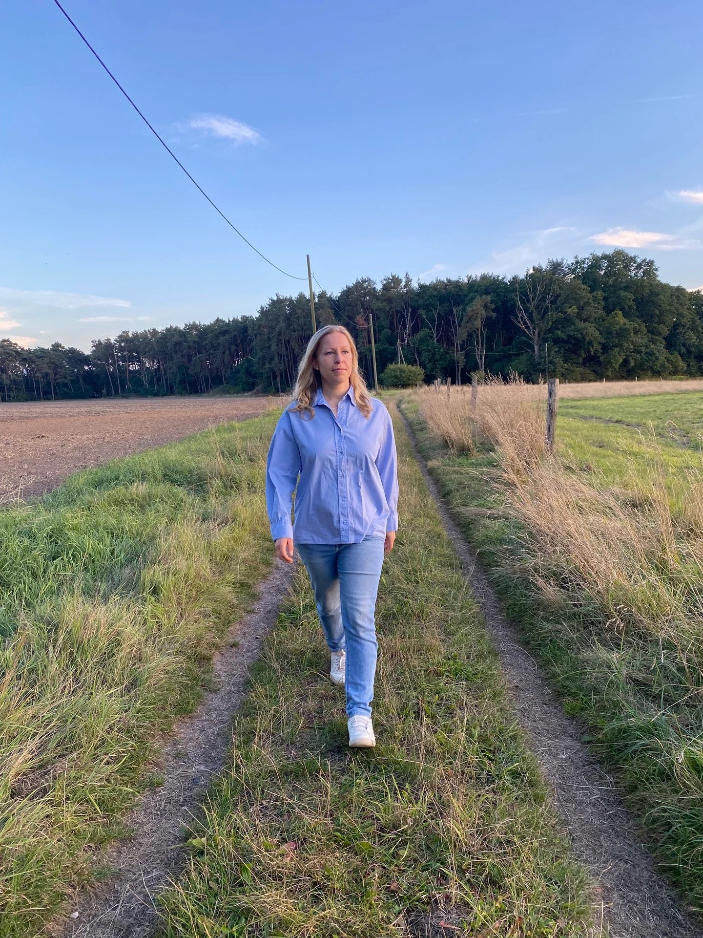 Eine Frau läuft auf einem schmalen Weg durch die Natur, im Hintergrund sind Felder, Bäume und ein blauer Himmel mit wenigen Wolken.