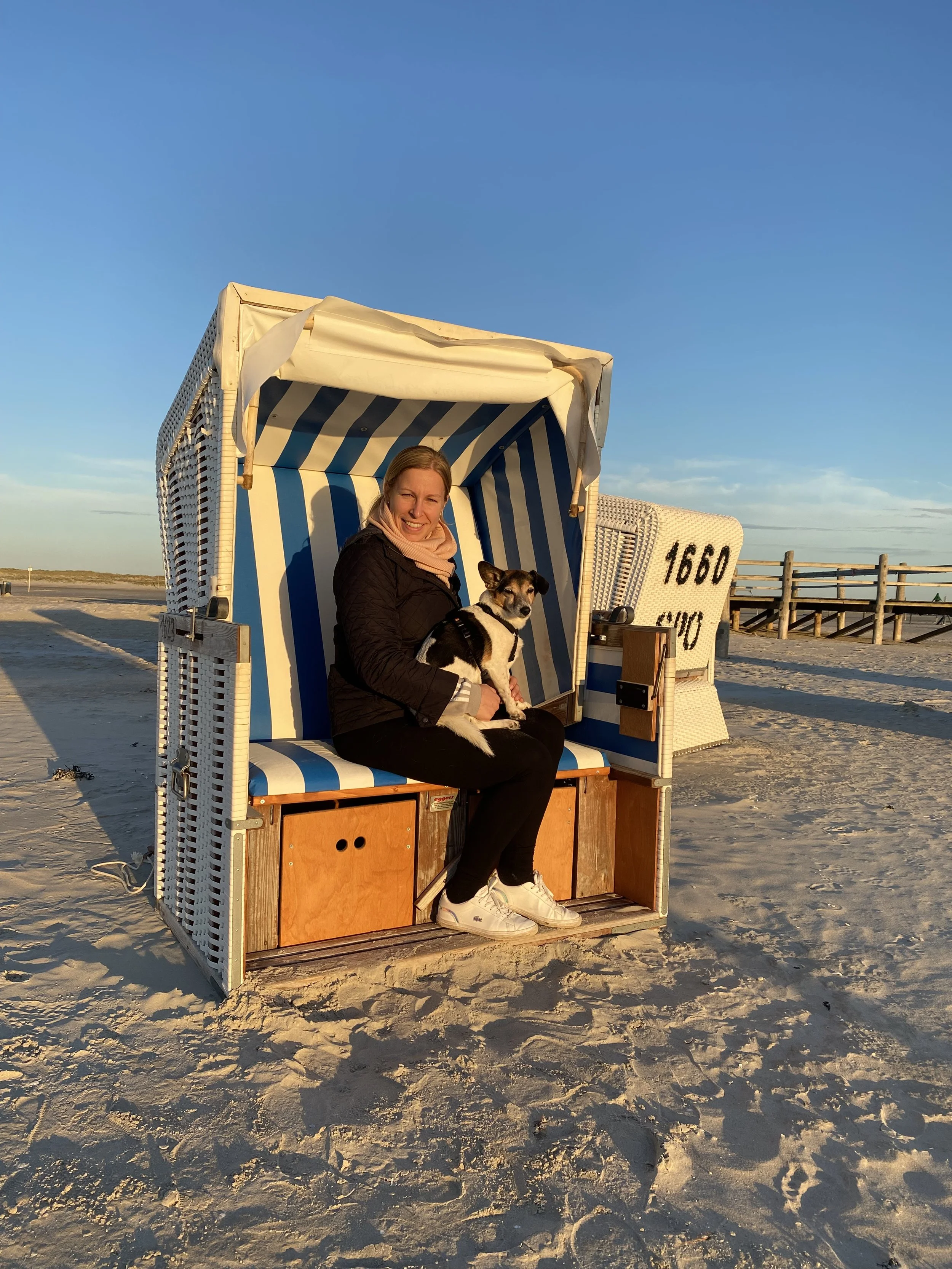 Eine Frau sitzt in einer Strandkörbchen mit einem Hund auf dem Sandstrand bei Sonnenuntergang, im Hintergrund ein Holzsteg und ein blauer Himmel.