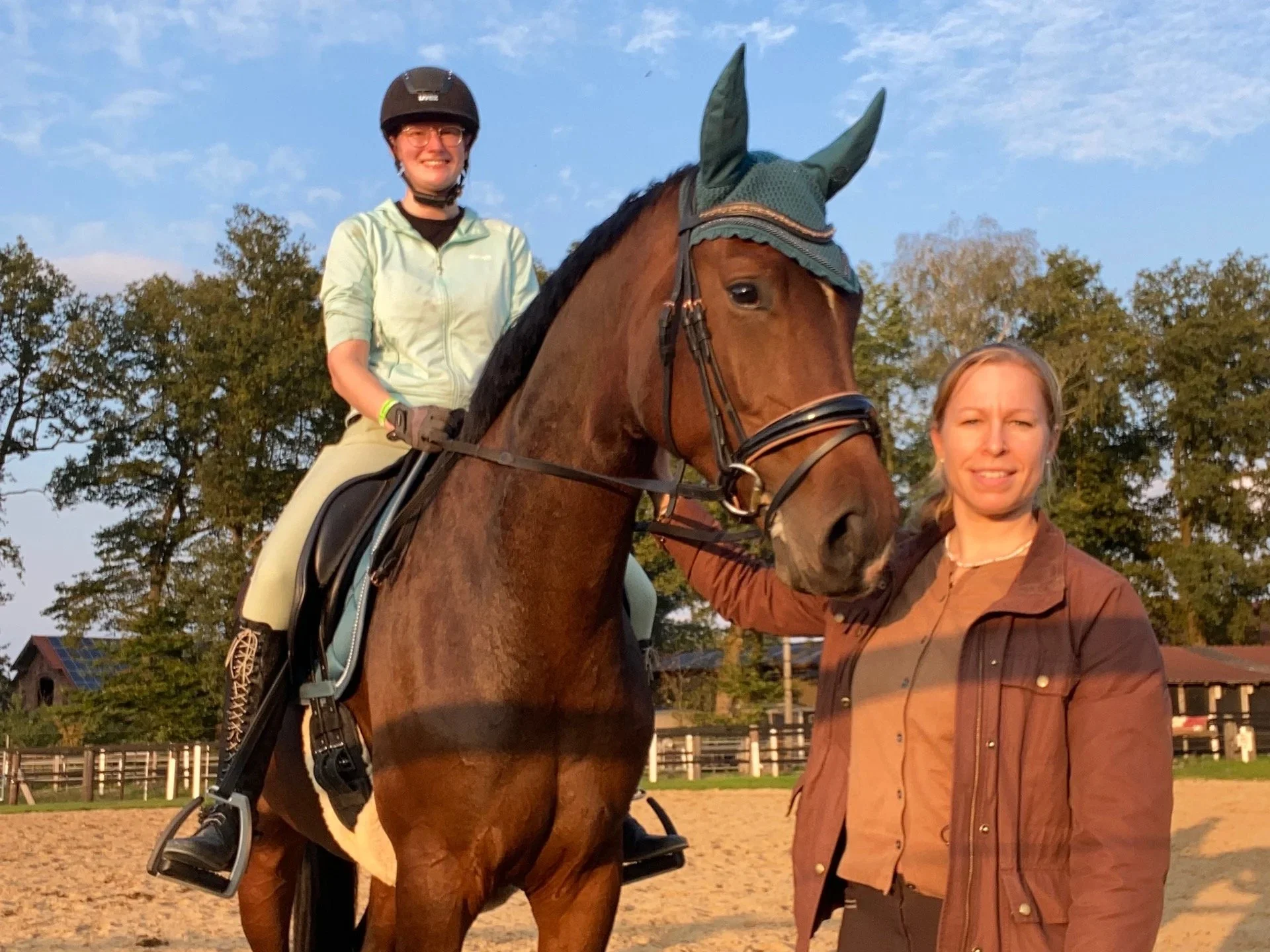 Zwei Frauen und ein braunes Pferd mit einem Reiter, der einen Helm trägt, in einem Reitplatz mit Bäumen im Hintergrund bei Sonnenuntergang.