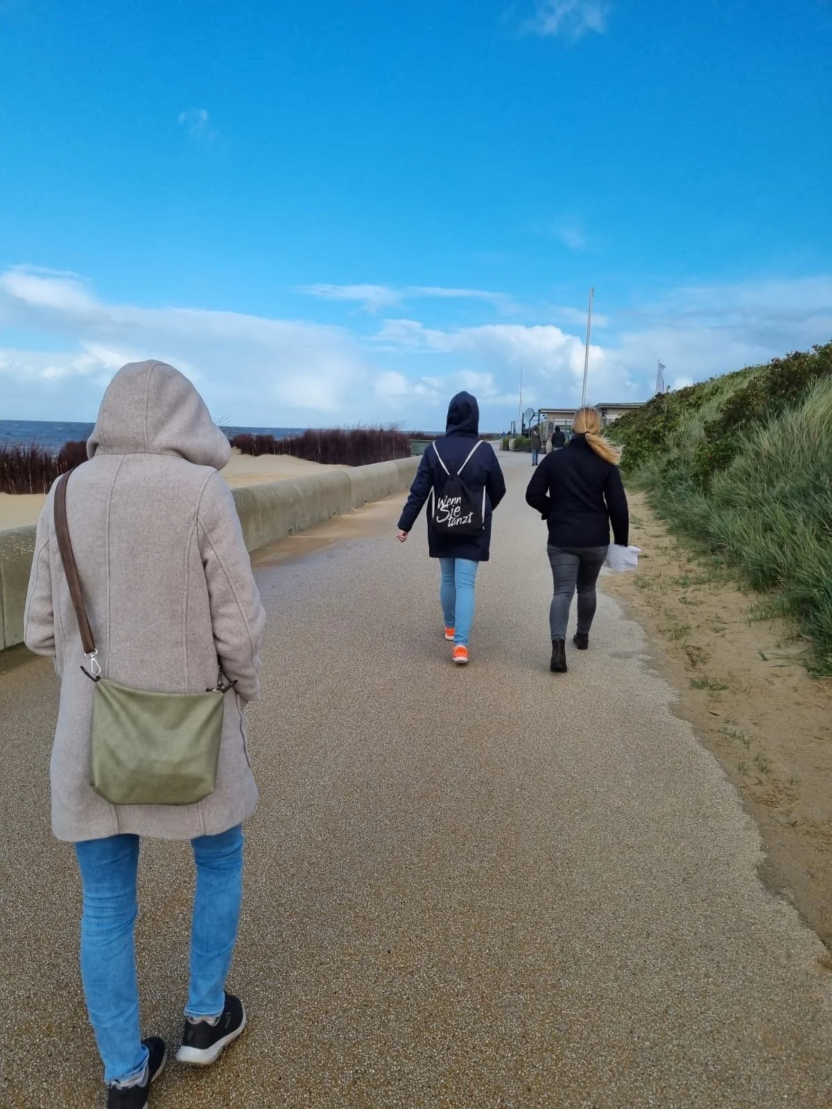 Drei Personen gehen an einem sonnigen Tag am Strand entlang, eine Person trägt einen grauen Pullover mit Kapuze, die anderen zwei tragen dunkle Jacken, die dritte hält eine weiße Tüte, im Hintergrund sind der Himmel, die Küste und grüne Büsche zu sehen.