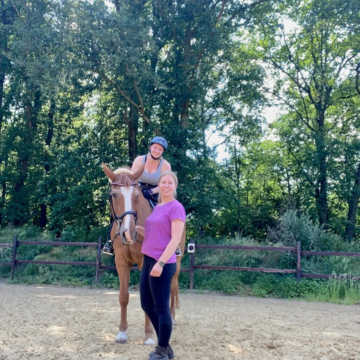 Zwei Frauen und ein Pferd in einem Reitstall im Freien, eine Frau sitzt auf dem Pferd und trägt einen Helm, die andere steht neben dem Pferd und lächelt.