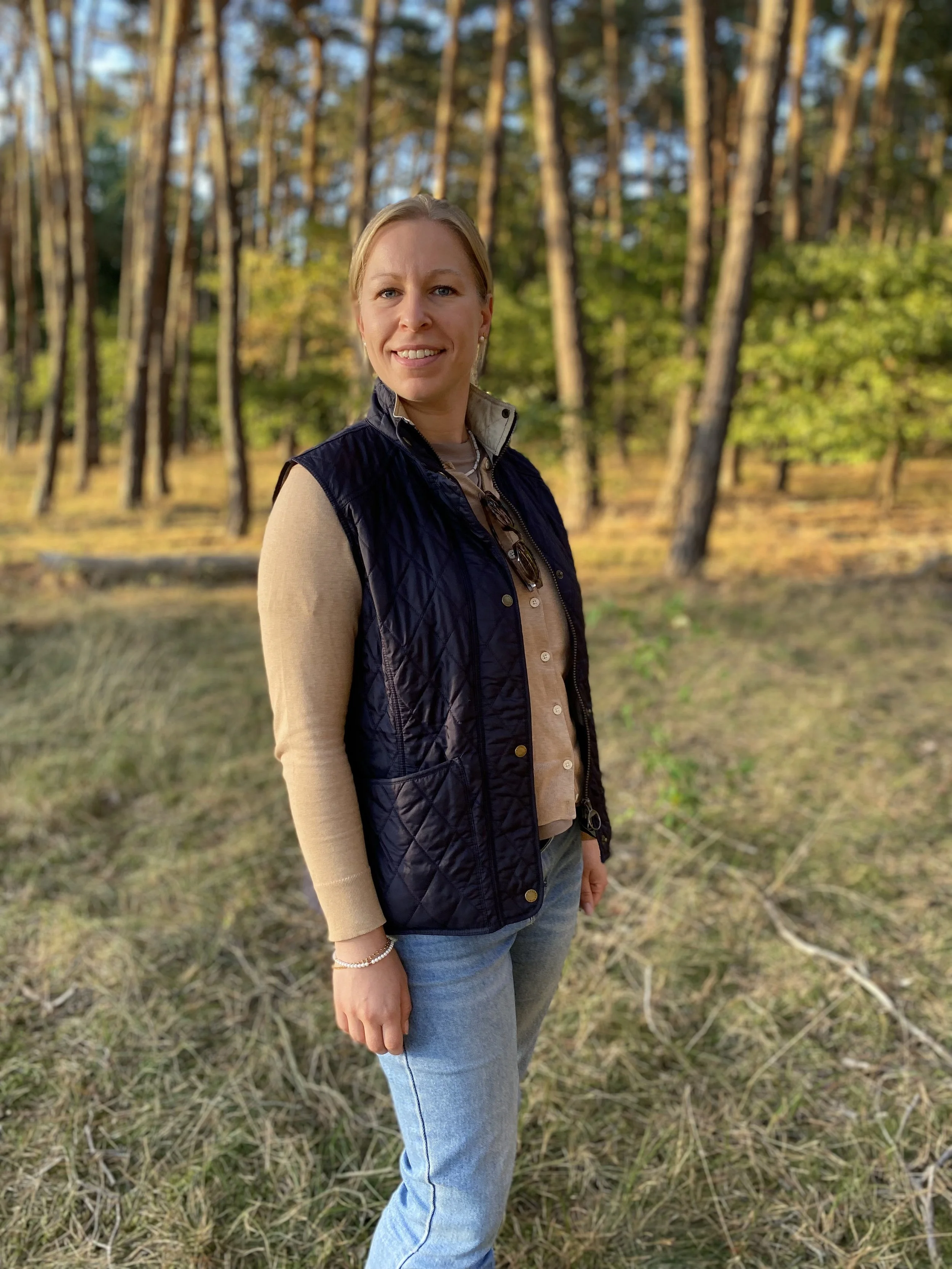 Eine Frau steht in einem Wald, trägt ein schwarzes gepunktes Oberteil, eine beige Weste und blaue Jeans. Es ist vermutlich spätsommerlich bis herbstlich.